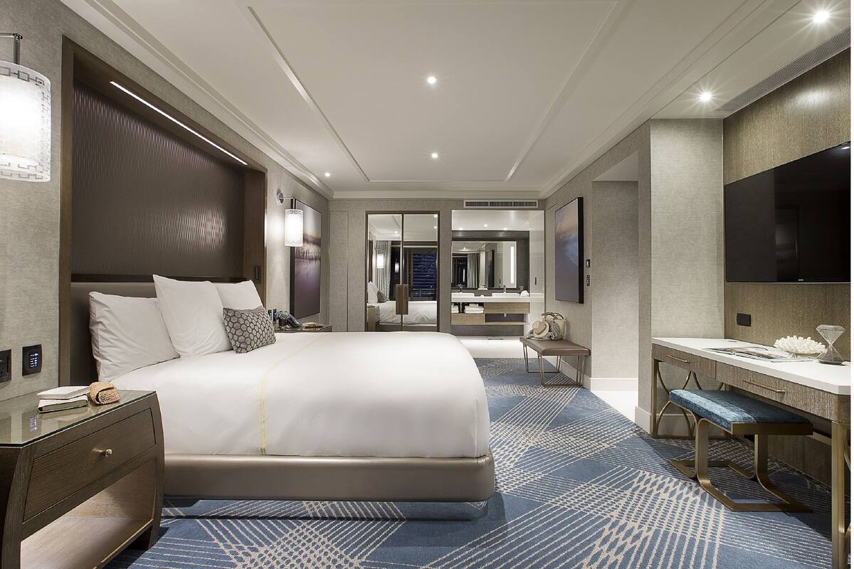 Une chambre d’hôtel moderne dotée d’un grand lit, d’un bureau avec une chaise, d’une télévision et d’une salle de bain visible à travers une porte en verre.