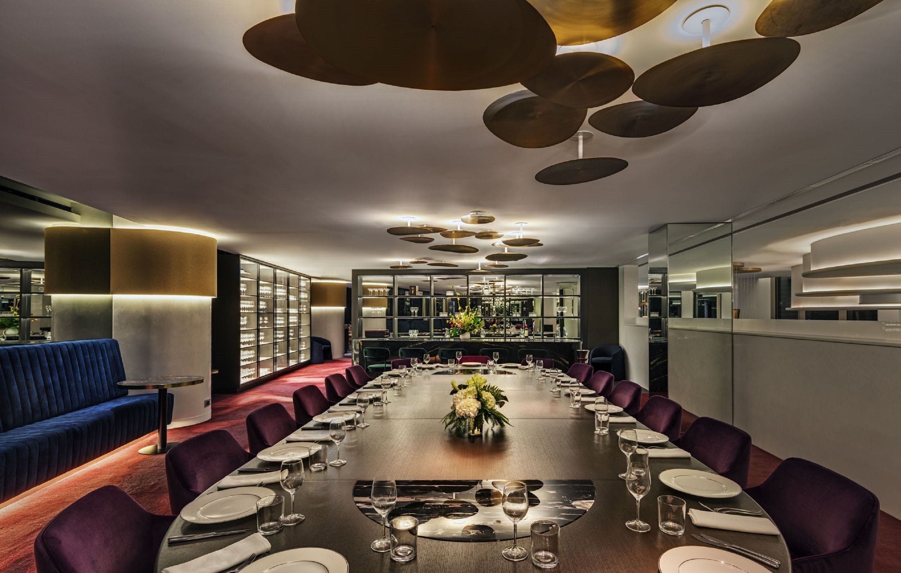 A modern dining area with a long table set for a meal, surrounded by purple chairs, and a blue sofa.