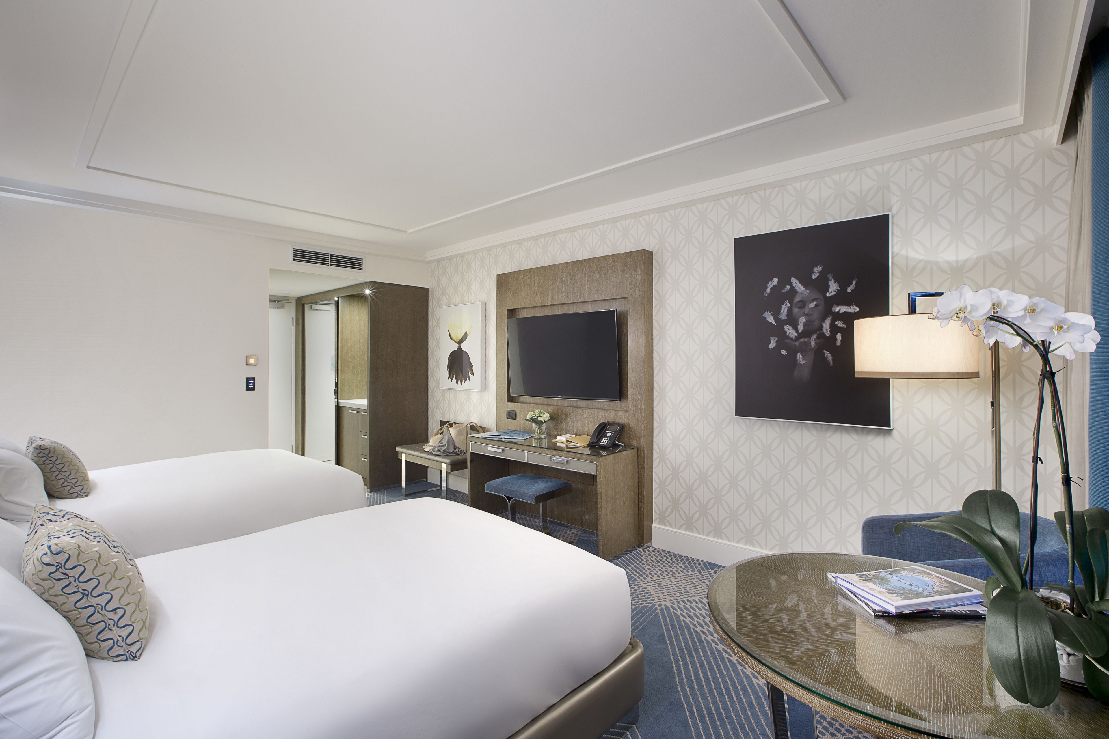Habitación de hotel con dos camas, un escritorio, televisión y zona de estar con lámpara y planta.
