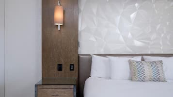 A hotel room with a wooden nightstand, a wall-mounted lamp, a bed with white bedding and patterned pillows, and a textured headboard.