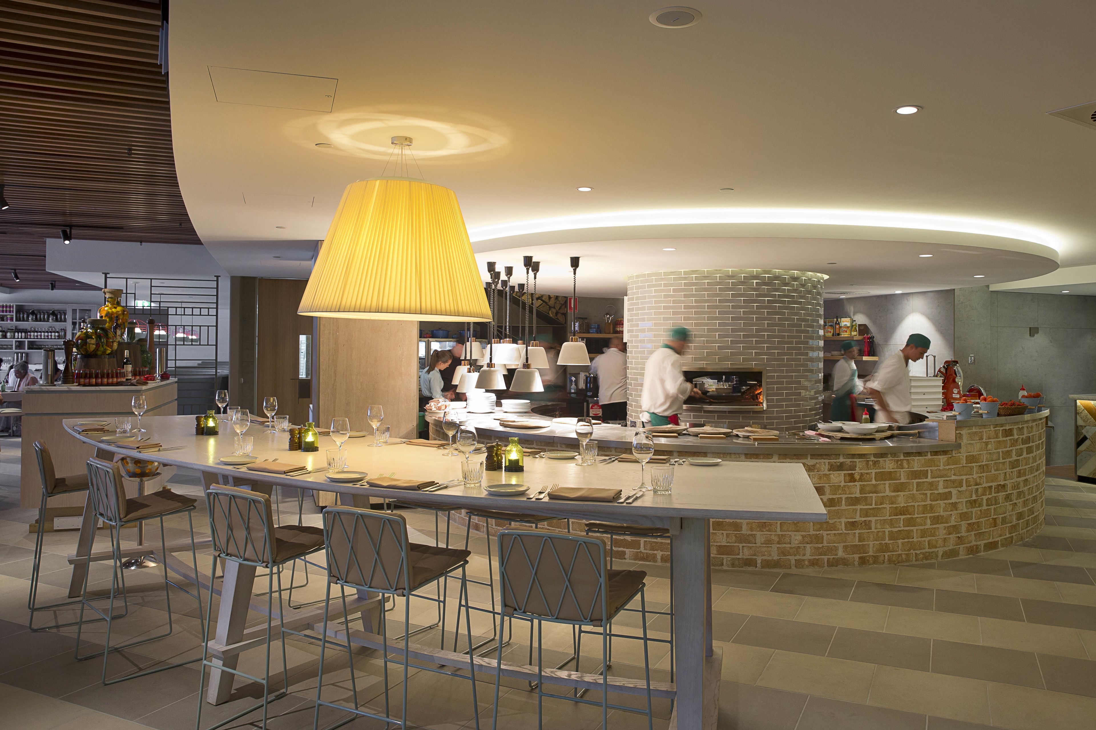 A modern restaurant with a central bar, seating area, and a visible kitchen.