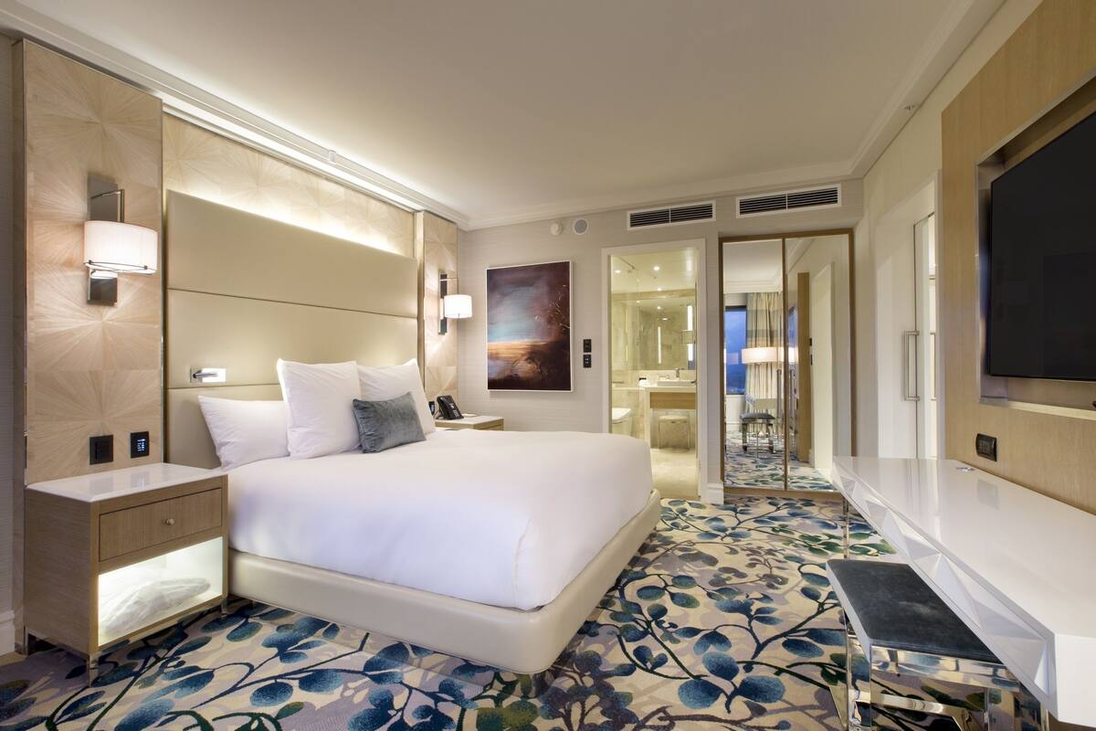 Une chambre d’hôtel moderne dotée d’un grand lit, de tables de chevet, d’un téléviseur à écran plat et d’une salle de bain visible à travers une porte ouverte.