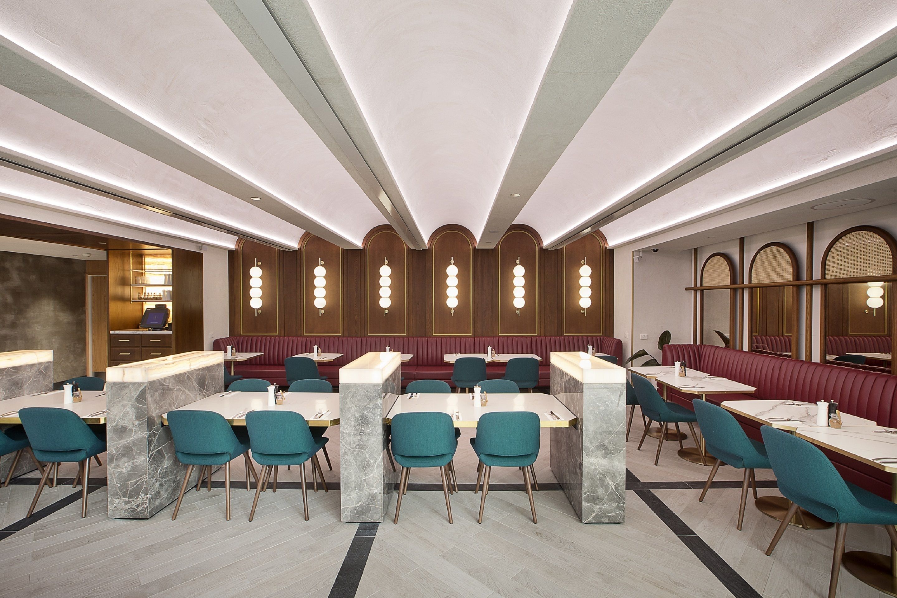 A modern restaurant with marble-topped tables and teal chairs, featuring a central seating area with red cushioned booths and a ceiling with linear lighting.