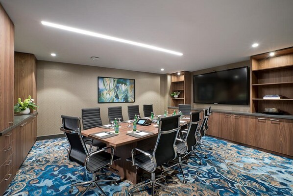 Una sala de conferencias con una gran mesa, sillas negras, un televisor de pantalla plana y un cuadro decorativo en la pared.