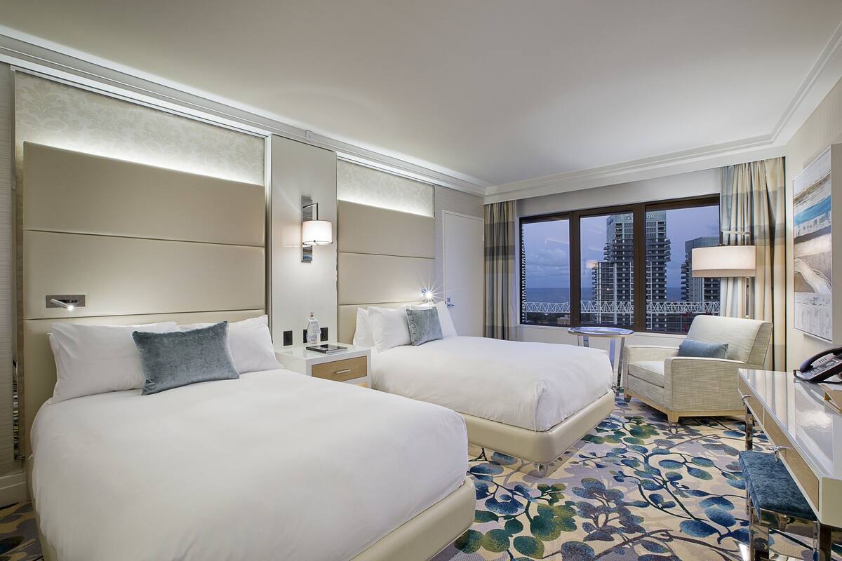 Une chambre d’hôtel avec deux lits, un coin salon, un bureau et une grande fenêtre donnant sur la ville.