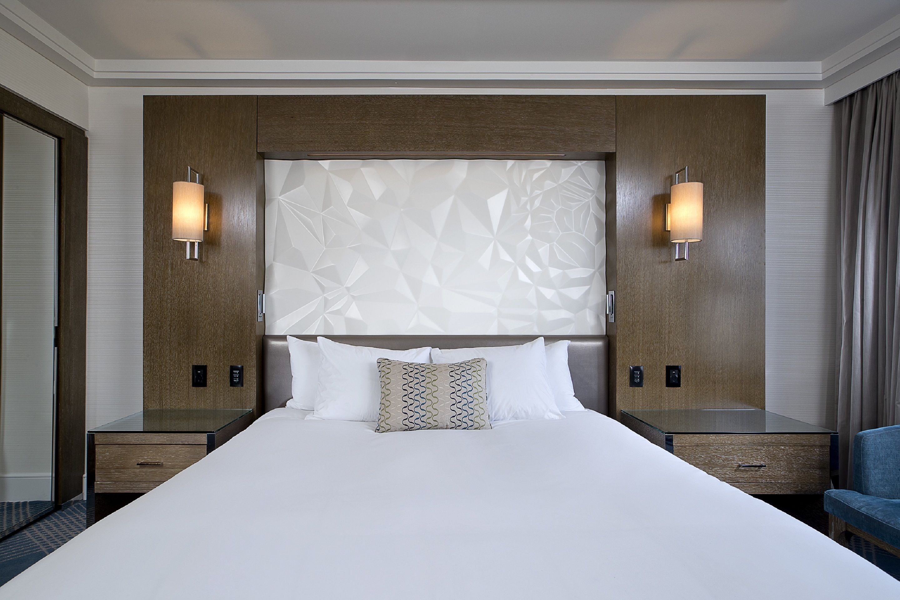 A hotel room with a large bed, two bedside tables, a nightstand, a blue armchair, and a geometric-patterned headboard.