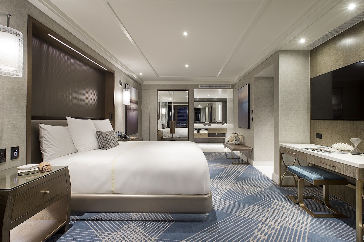 Habitación de hotel moderna con una cama grande, un escritorio, un televisor de pantalla plana y un baño visible a través de una puerta de vidrio.