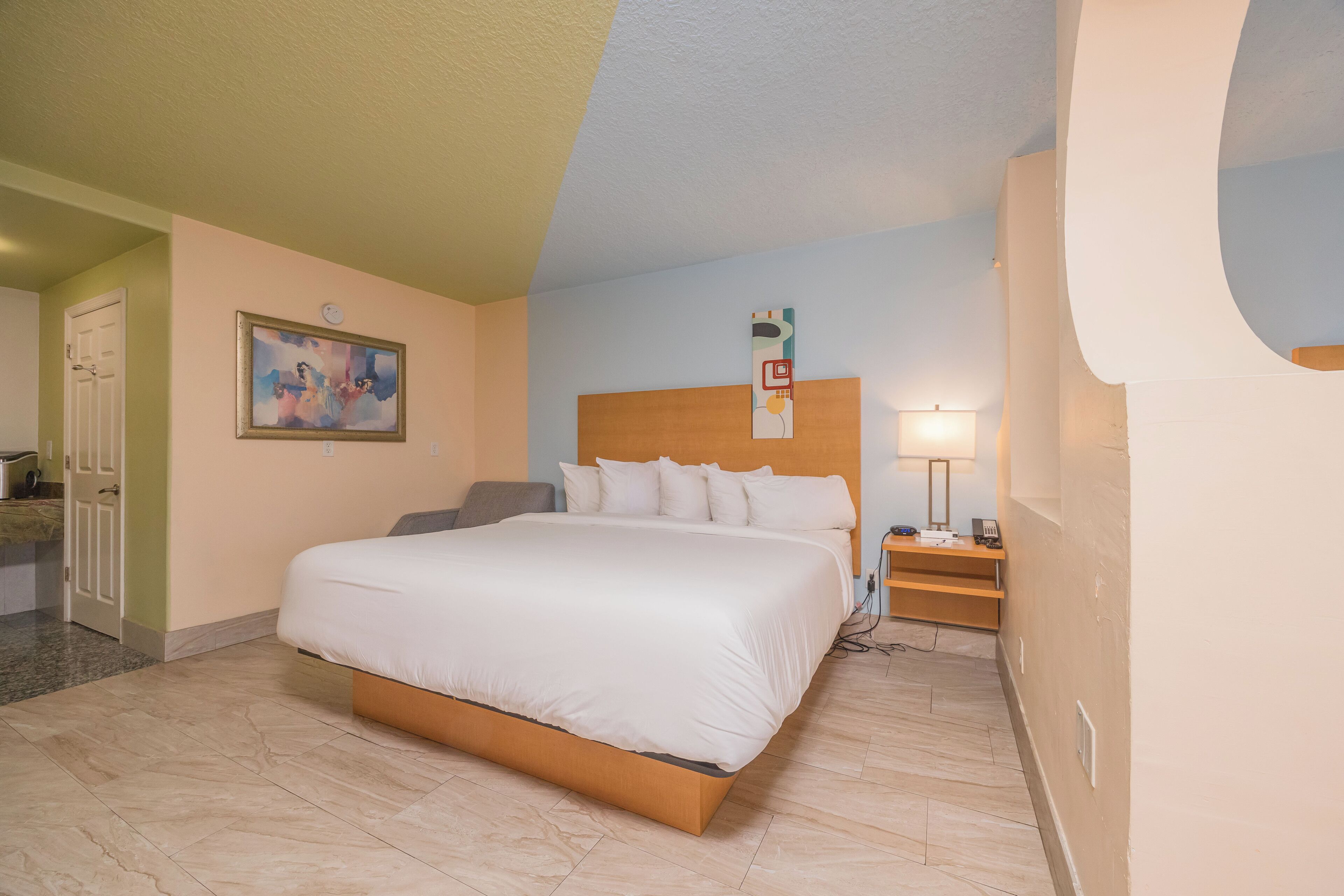 A hotel room with a large bed, a bedside table with a lamp, a painting on the wall, and a door to the left.
