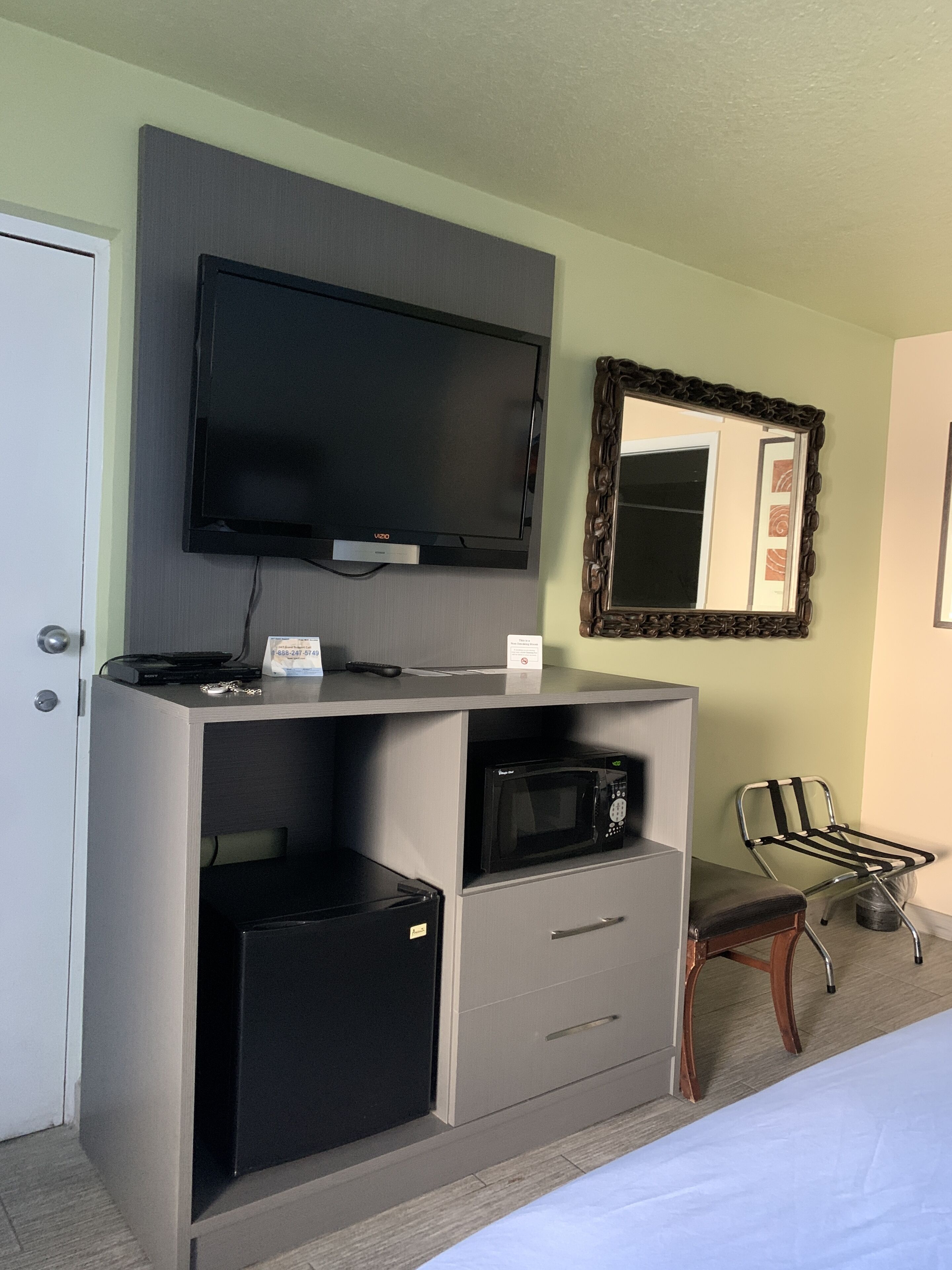 A hotel room with a flat-screen TV mounted on a grey cabinet, a microwave, a small refrigerator, a chair, and a bed.
