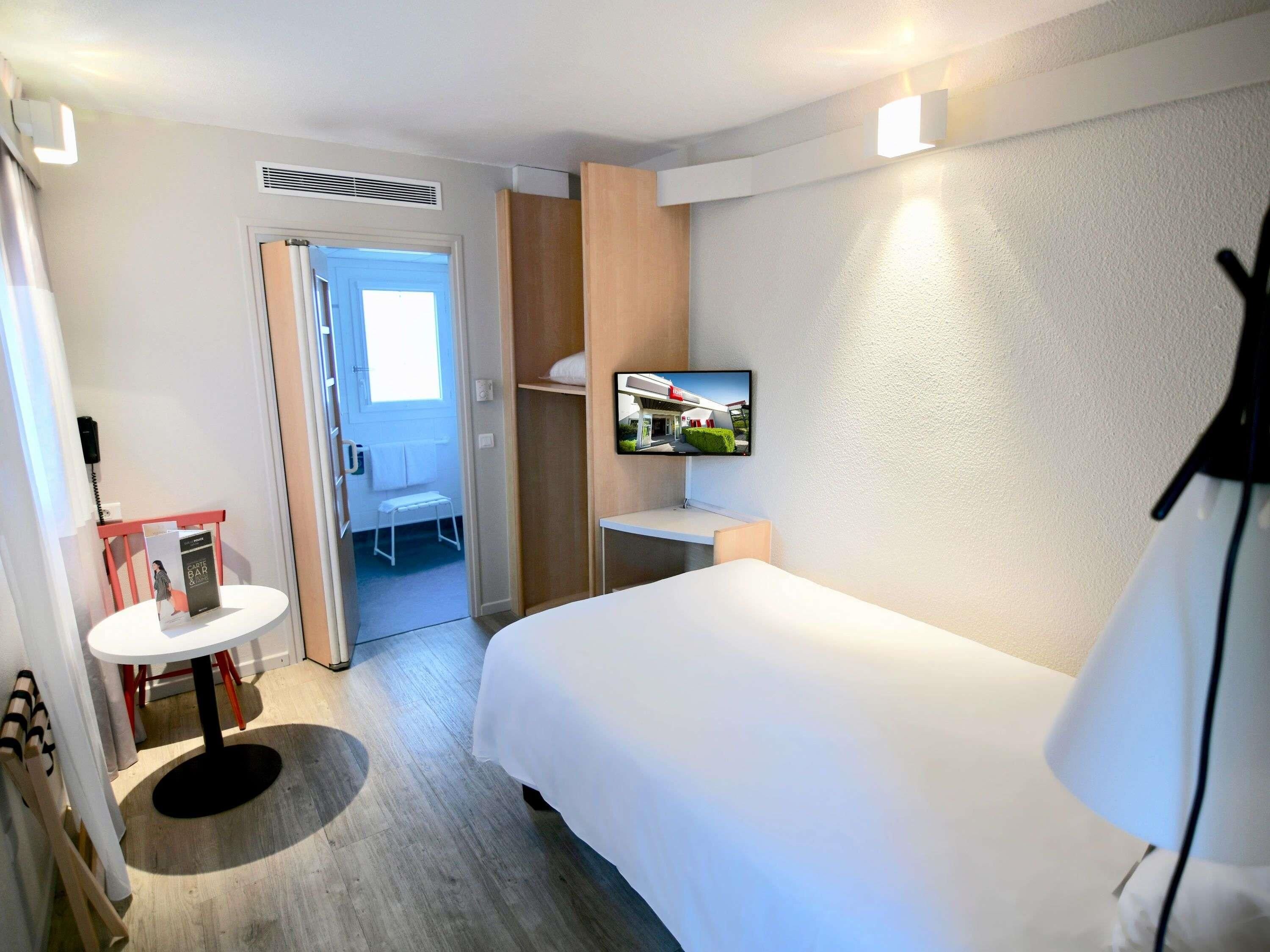 Quarto de hotel com uma cama, televisão presa na parede, uma mesinha e uma cadeira.