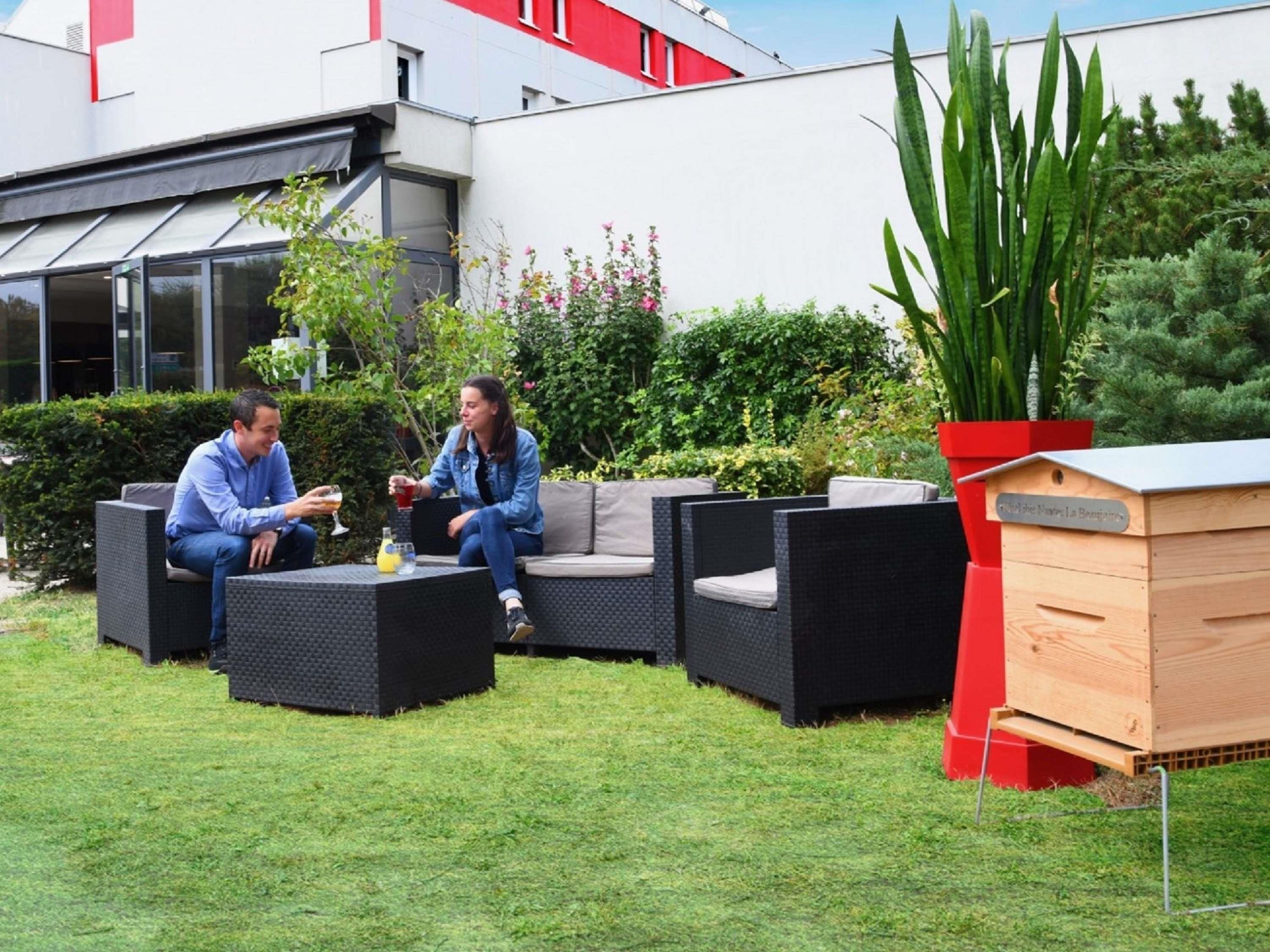 Two people seated on outdoor furniture in a garden area with a beehive and a building in the background.