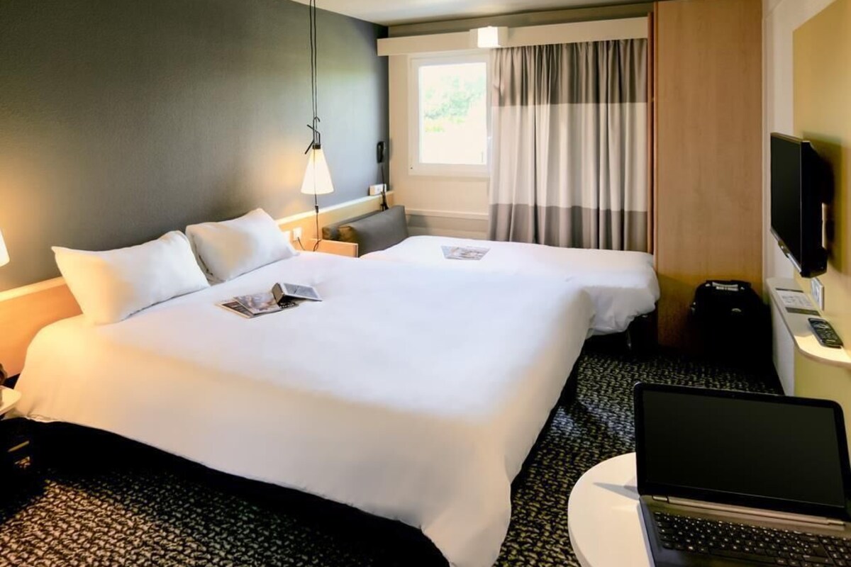 Habitación de hotel con dos camas, un escritorio con una computadora portátil, un televisor y una ventana con cortinas.