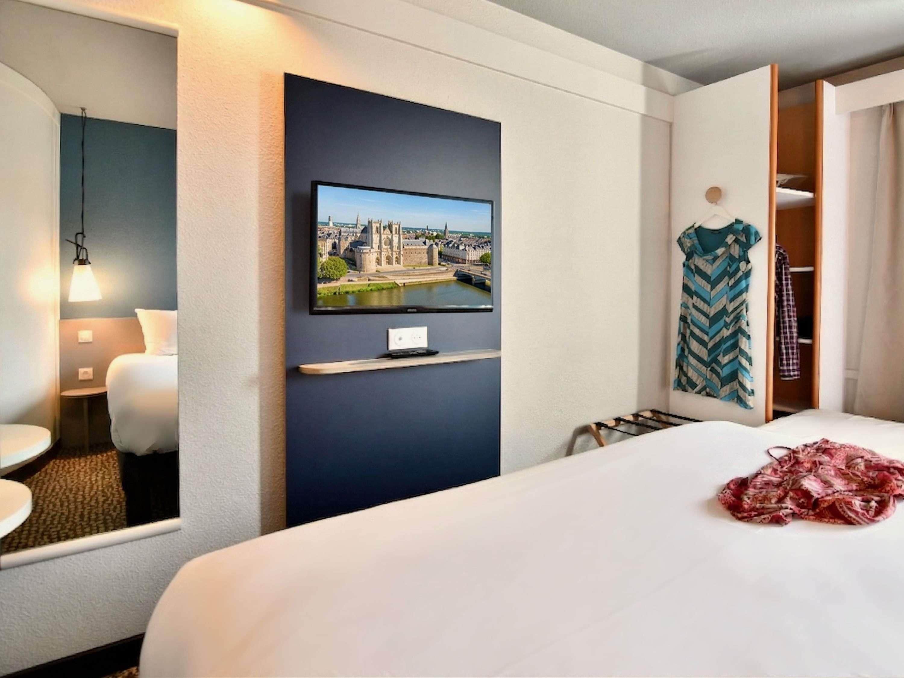 A hotel room with a bed, a television mounted on the wall, a mirror, a bedside table, and a wardrobe.