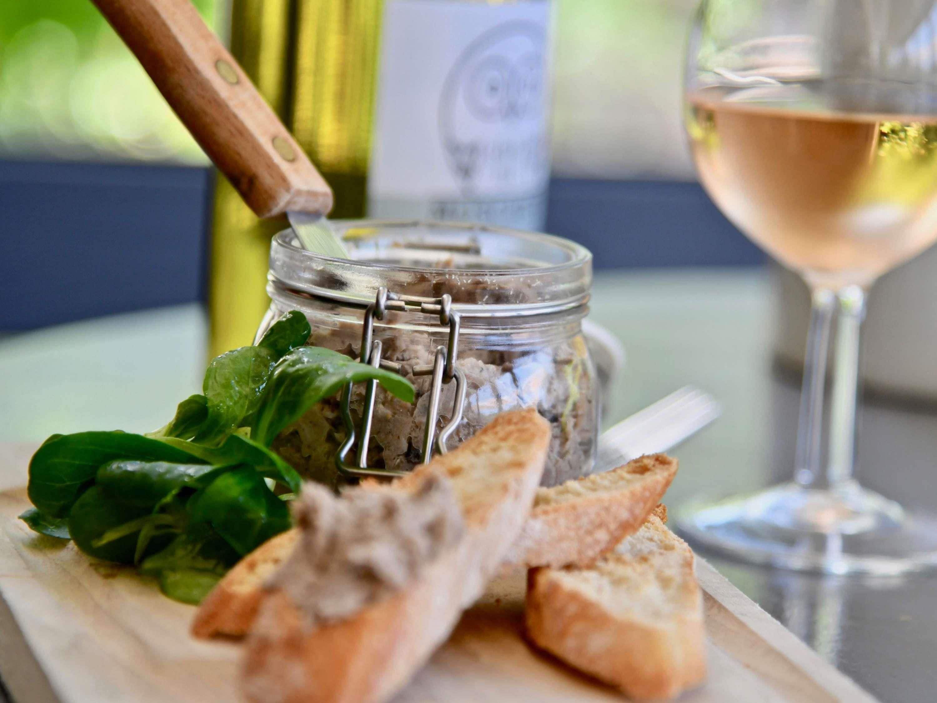 A wooden cutting board with a jar of spread, some bread, and a glass of wine.
