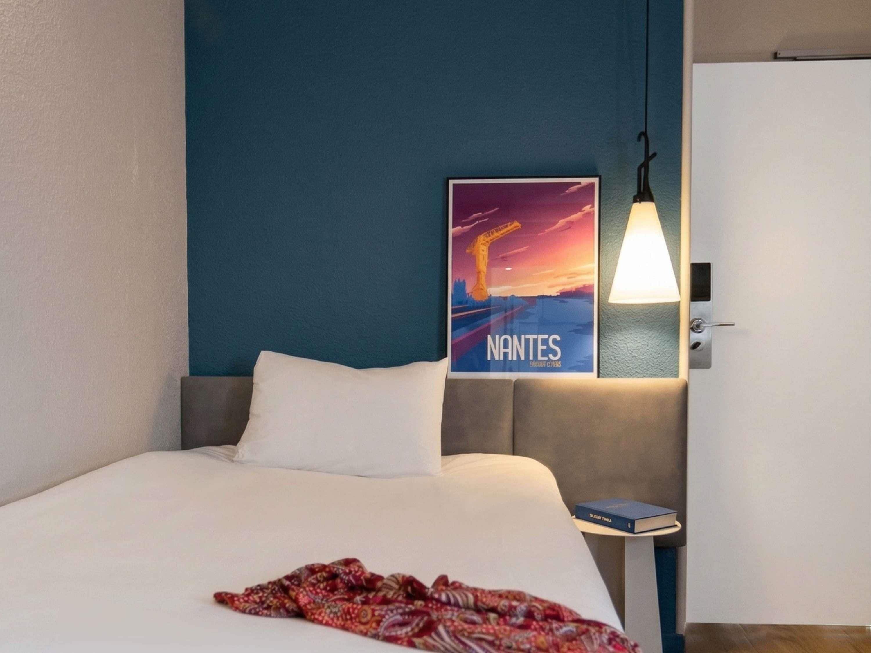 Une chambre d’hôtel avec un lit, une table de chevet sur laquelle se trouve un livre, une affiche au mur et un luminaire suspendu.
