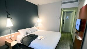 A hotel room with a neatly made bed, bedside lamps, a TV on the wall, and a small table with a chair. - ibis Nantes La Beaujoire Parc Expo (Nantes)