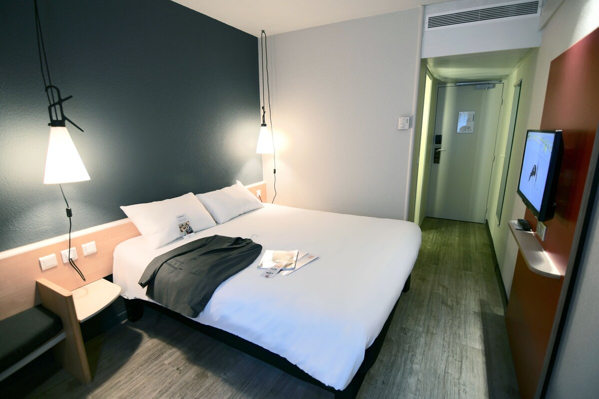 Habitación de hotel con una cama bien hecha, lámparas de noche, un televisor en la pared y una mesita con una silla.