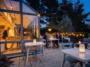 Un espace extérieur aménagé pour les repas, avec des tables et des chaises, une véranda en verre offrant une vue sur l’intérieur, et des guirlandes lumineuses en arrière-plan.