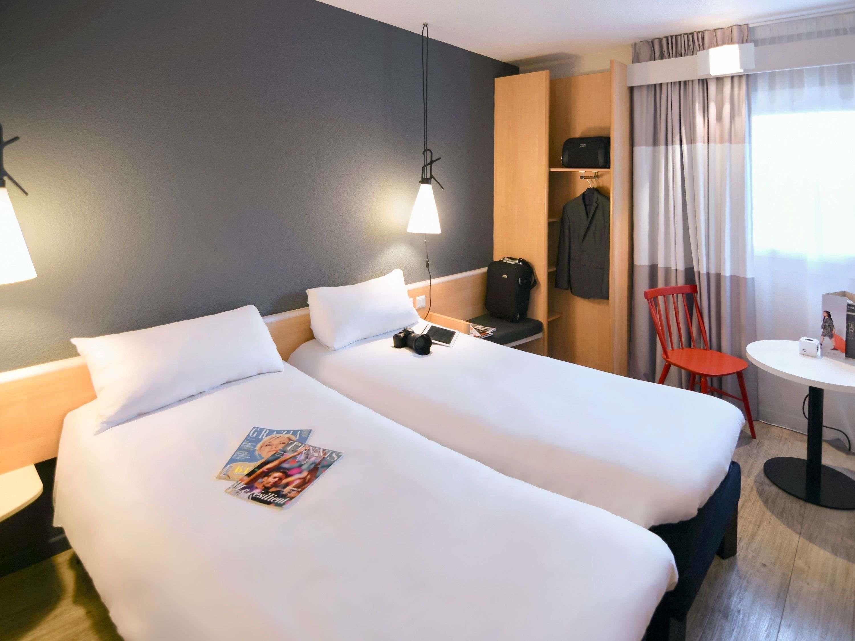 Habitación de hotel con dos camas, un escritorio, una silla, una mesa, un armario y una ventana con cortinas.