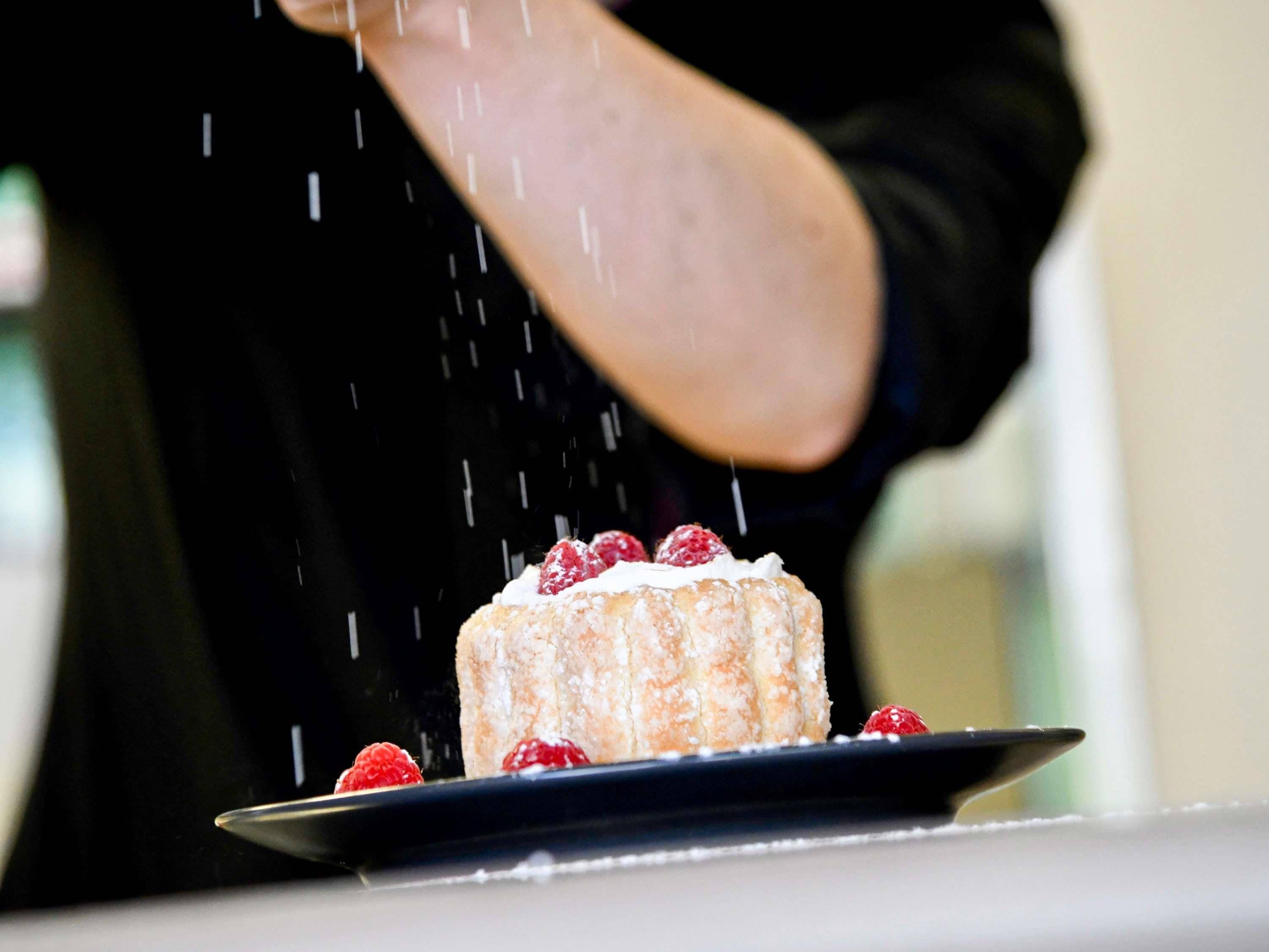 Une personne saupoudre du sucre glace sur un gâteau décoré de framboises, le tout présenté sur une assiette.