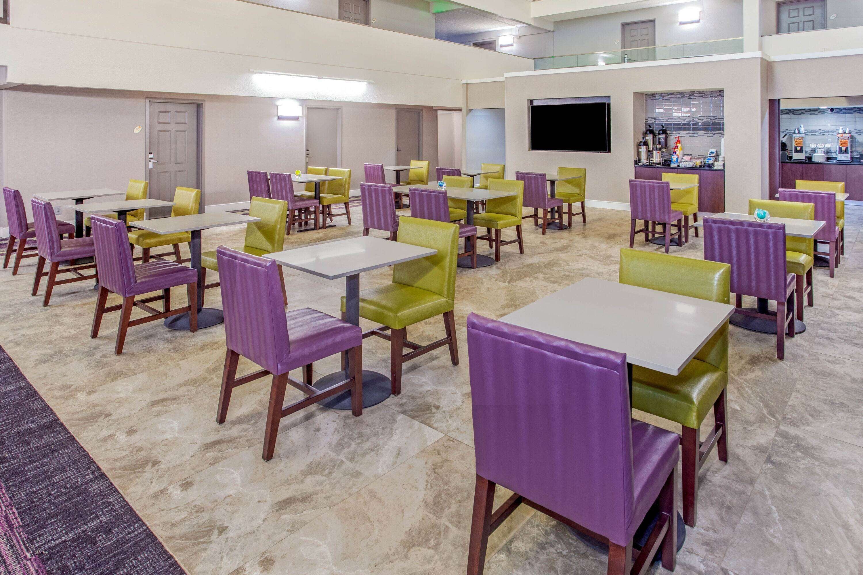 A hotel dining area with multiple tables and chairs, a flat-screen TV, and a bar counter with various items.