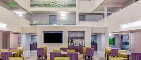 A modern dining area with multiple tables and chairs, a flat-screen TV mounted on the wall, and a kitchen area in the background.