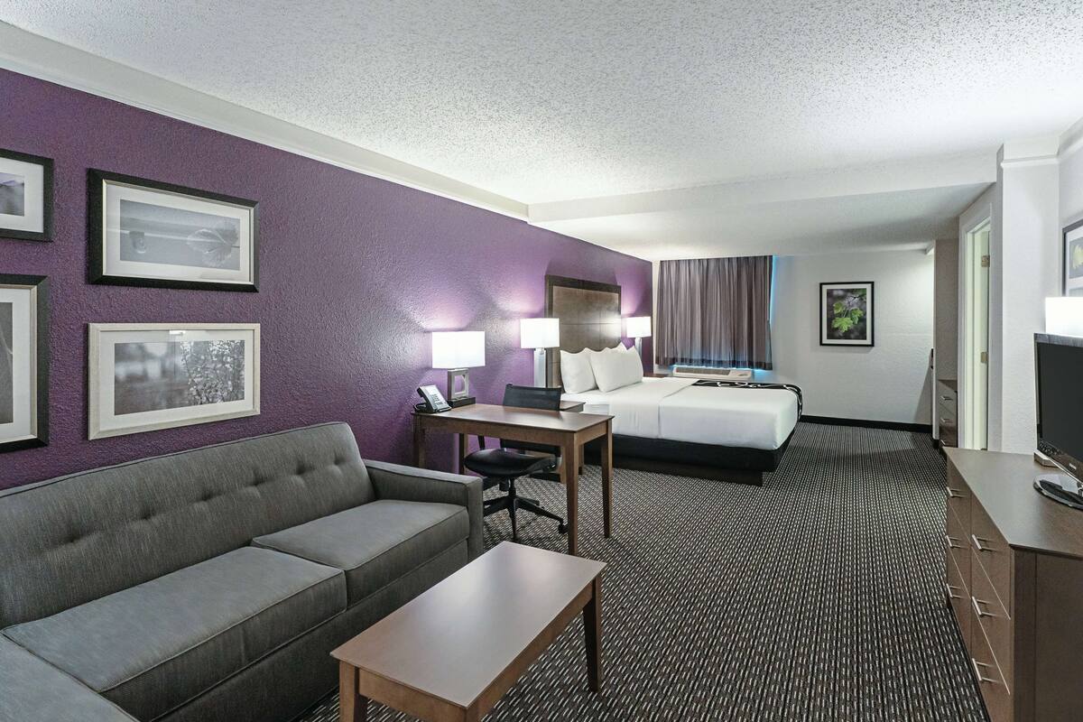 A hotel room with a grey sofa, a wooden table, a bed with white linens, and a television.