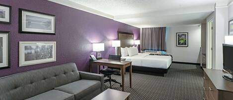 A hotel room with a grey sofa, a wooden table, a bed with white linens, and a television.