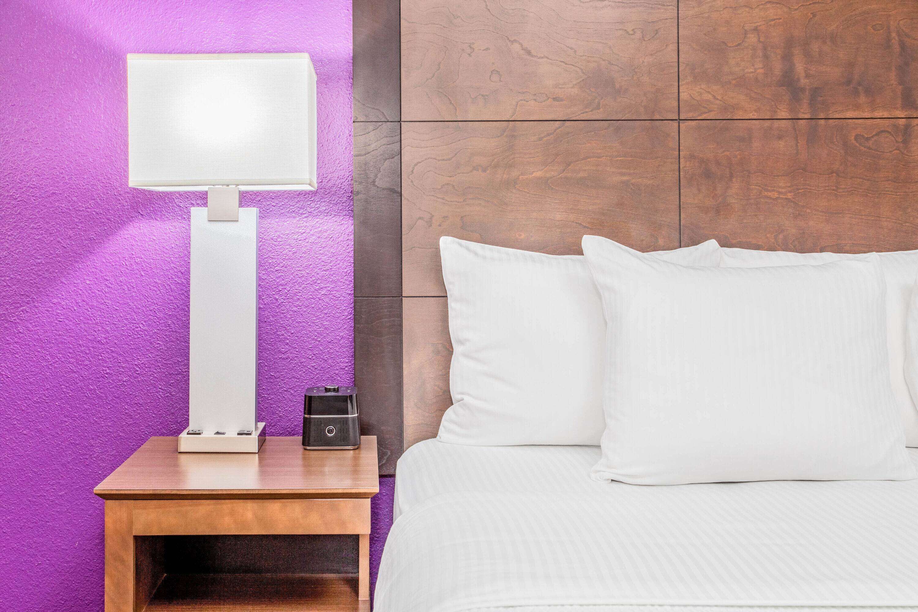 A neatly made bed with white bedding, a bedside table with a lamp, and a wall with a purple texture.