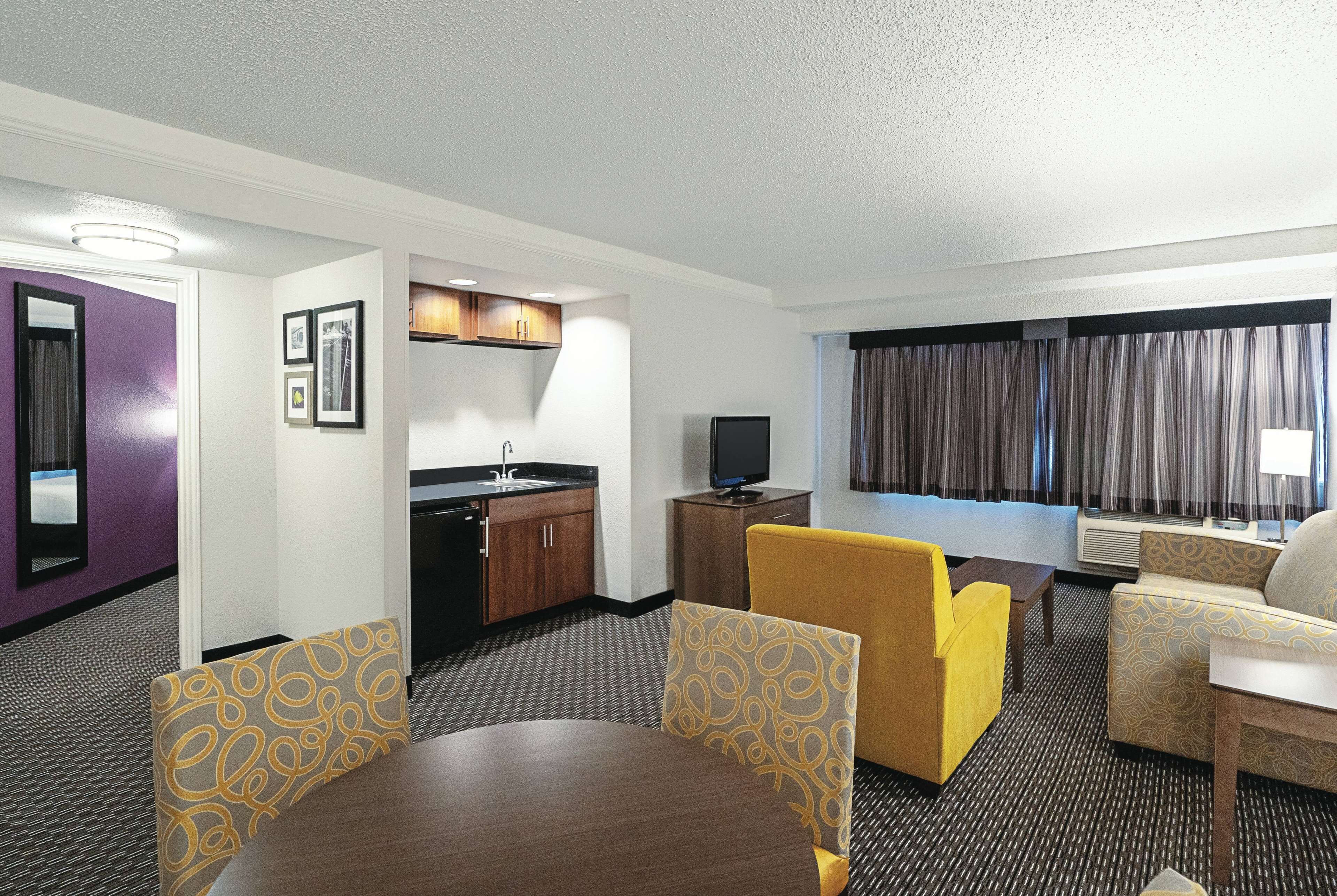 A hotel room with a dining table, yellow armchairs, a sofa, a television, a kitchenette, and a large window with curtains.
