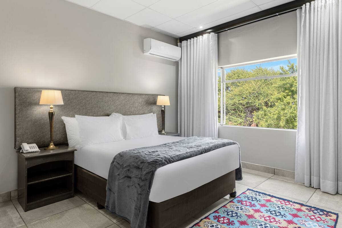 Une chambre d’hôtel avec un lit, deux lampes de chevet, un téléphone, une fenêtre avec des rideaux et un tapis à motifs.