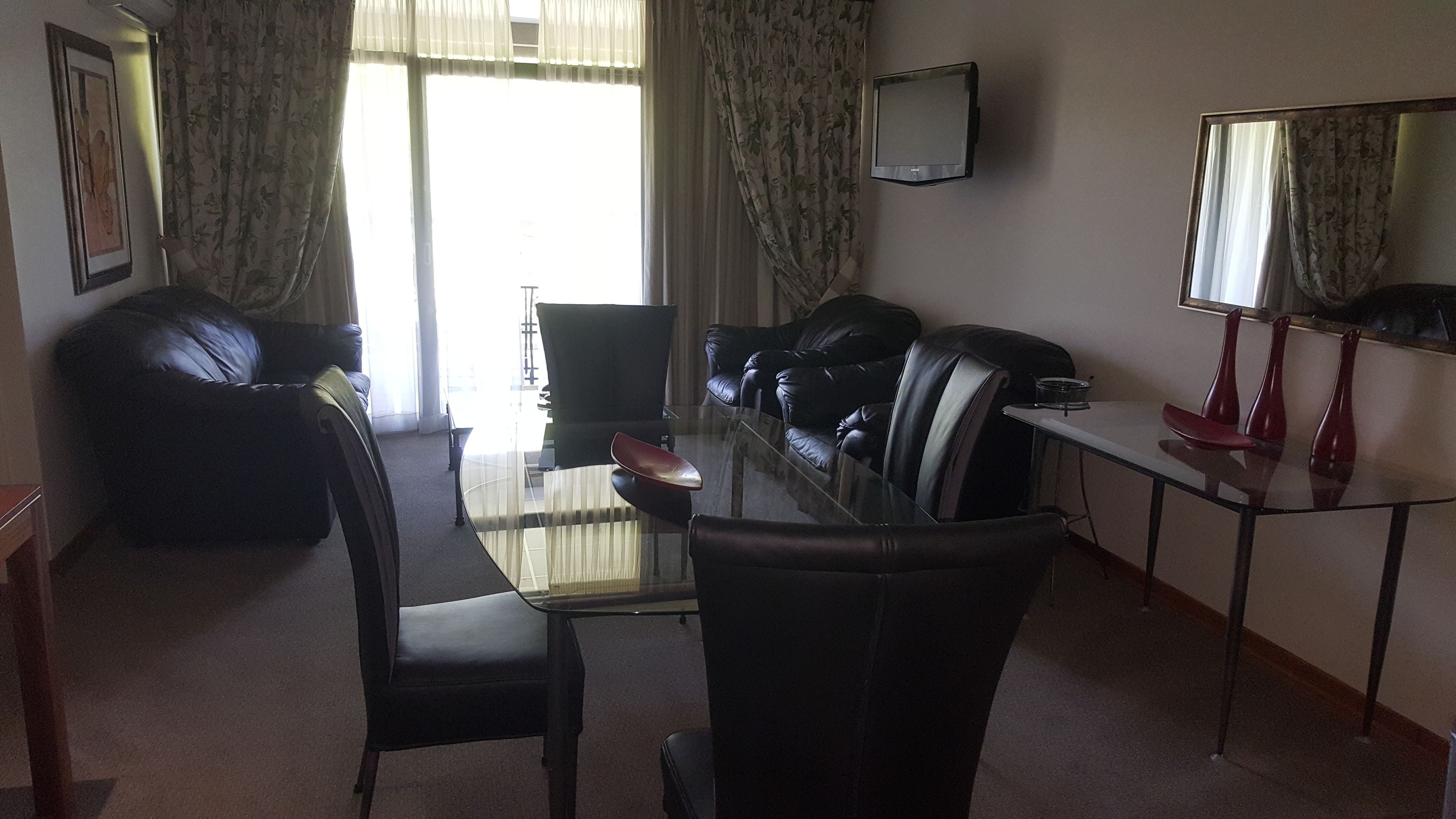 A living room with a glass coffee table, leather sofas, a TV mounted on the wall, and a dining area with a small table and chairs.