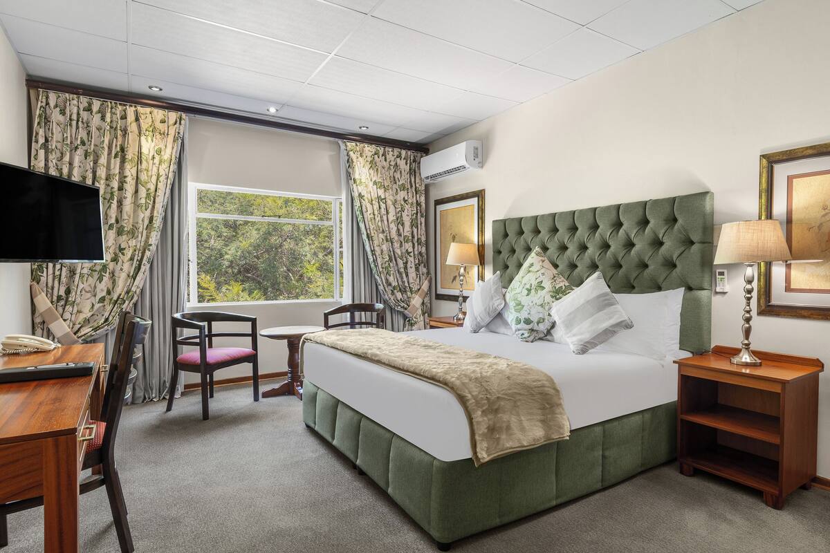 Une chambre d’hôtel avec un lit recouvert d’un tissu vert, un téléviseur à écran plat, un bureau avec une chaise, une fenêtre avec des rideaux et une table de chevet avec une lampe.