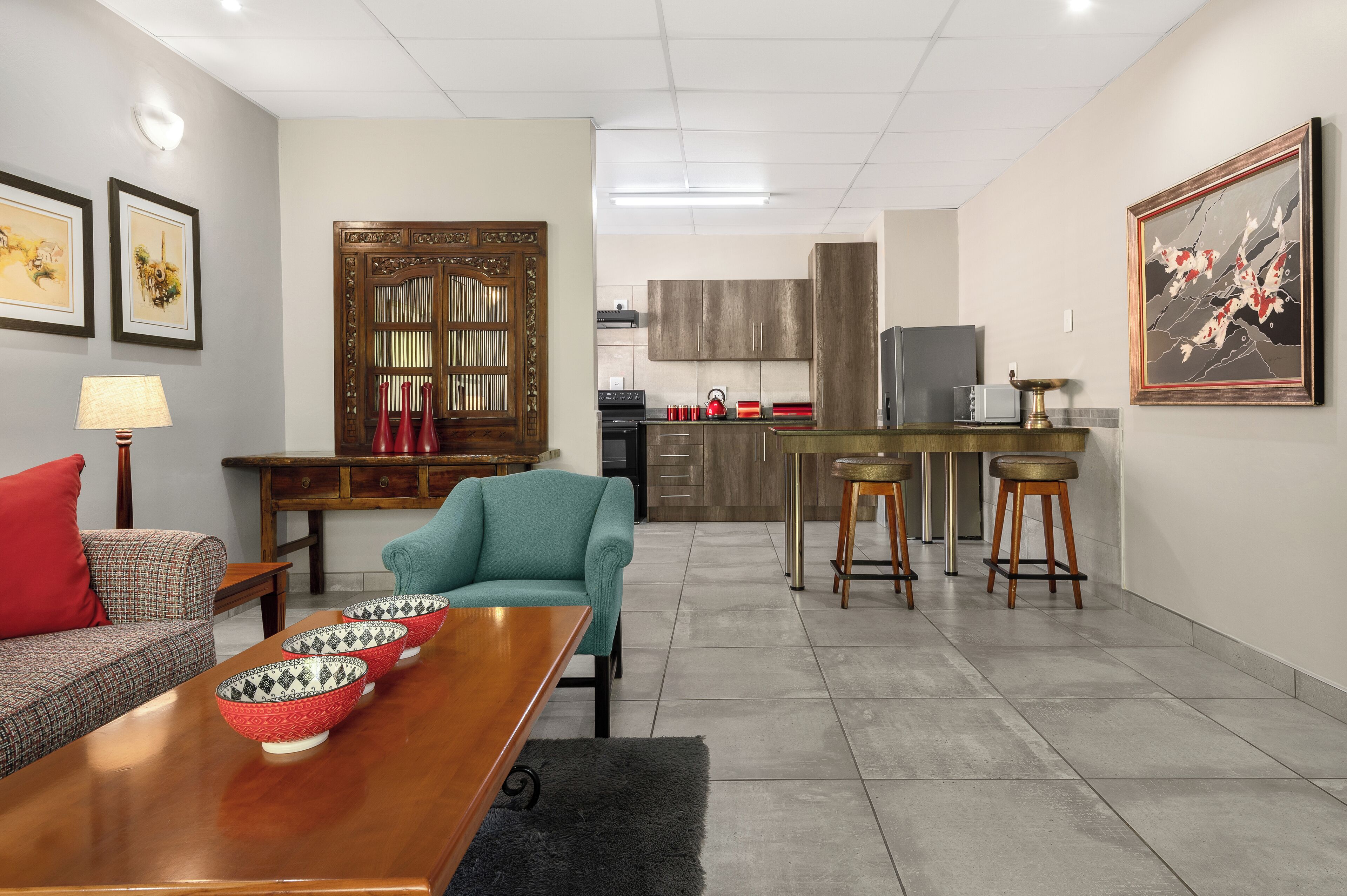 A modern living room with a sofa, a teal armchair, a wooden coffee table with decorative bowls, a dining table, and a kitchen in the background.