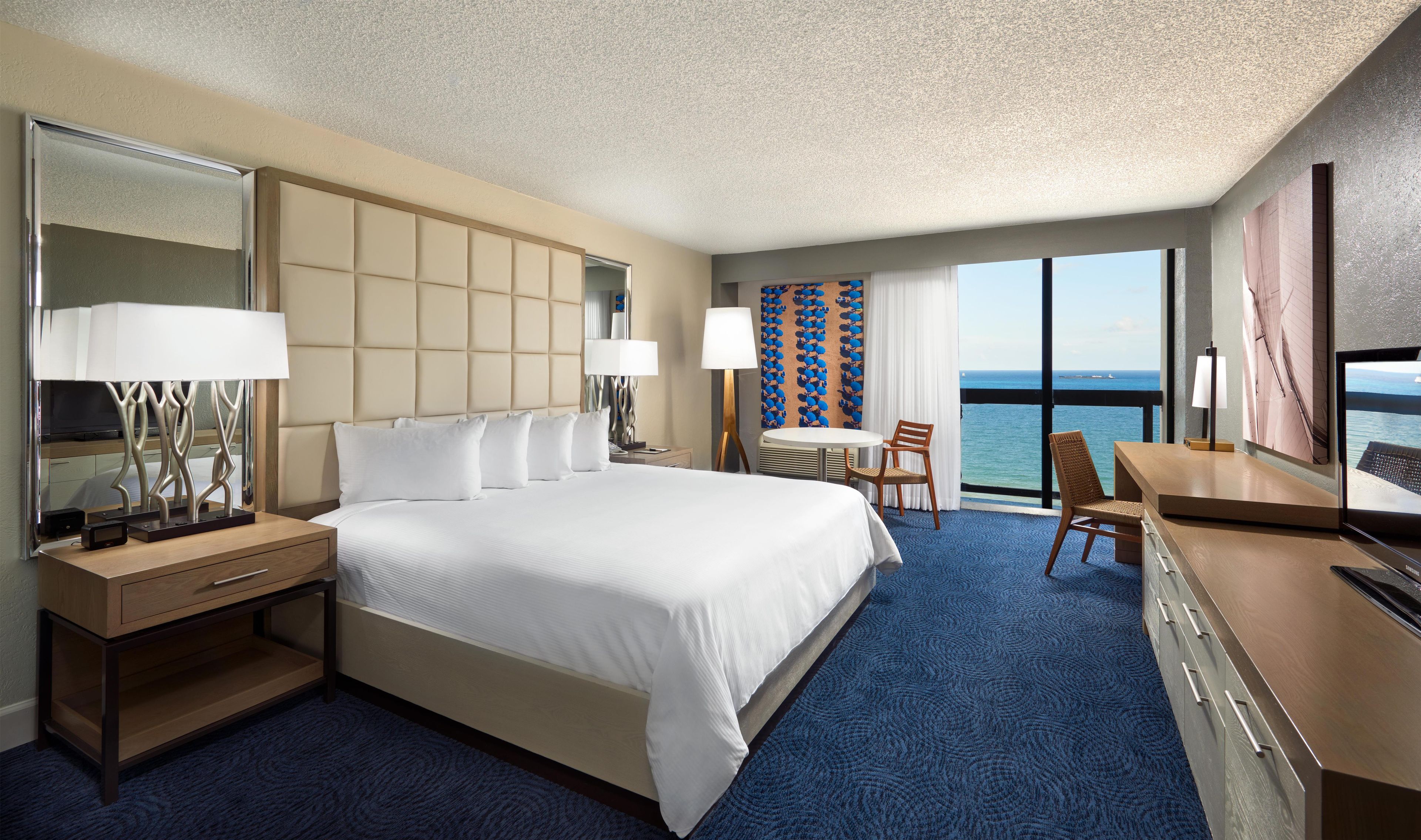 A hotel room with a large bed, a desk, a chair, a television, and a view of the sea.