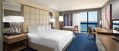 A hotel room with a large bed, a desk, a chair, a television, and a view of the sea.