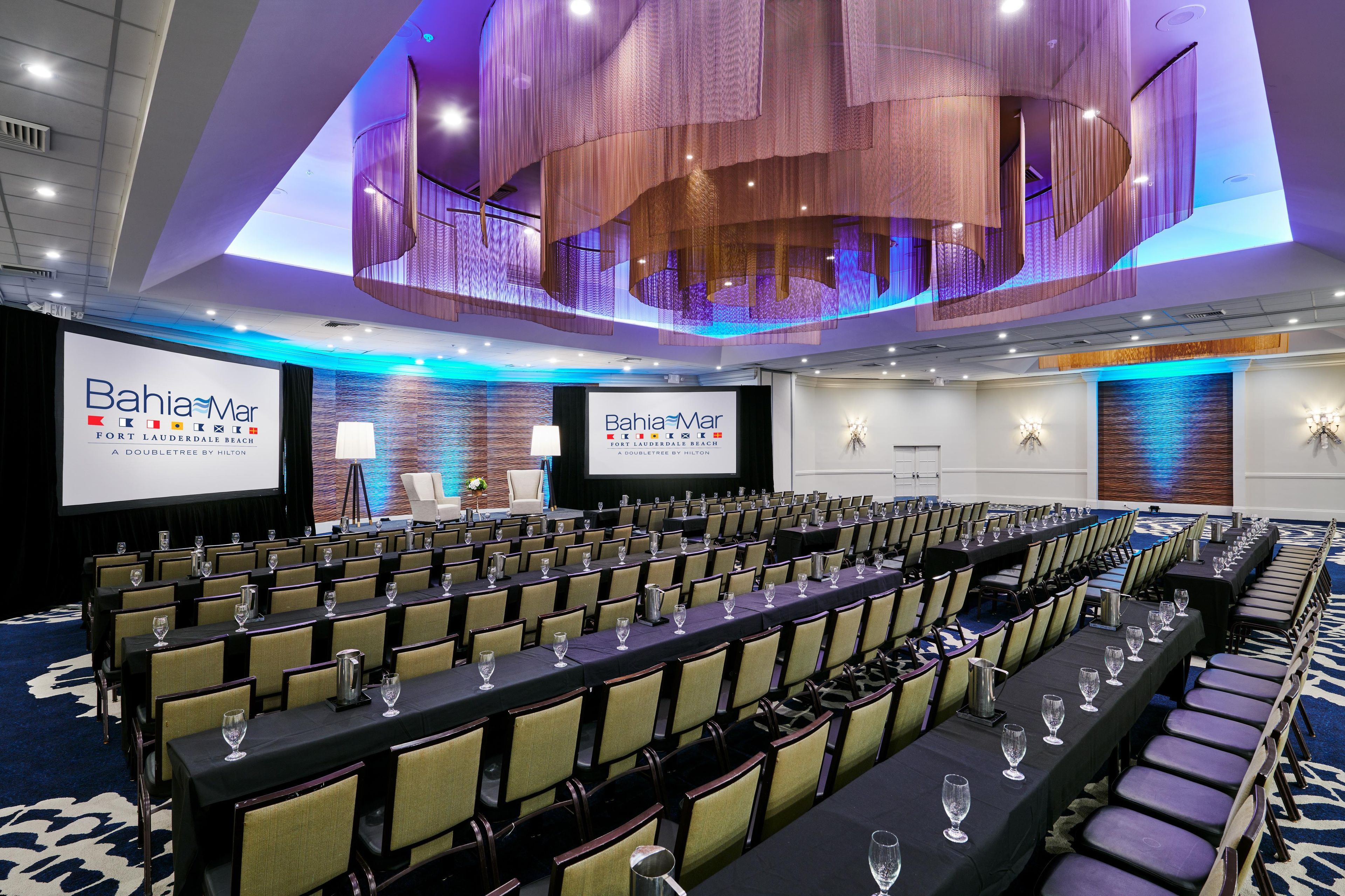 A conference room with rows of tables and chairs, a large screen displaying the BahiaMar logo, and a modern ceiling design.