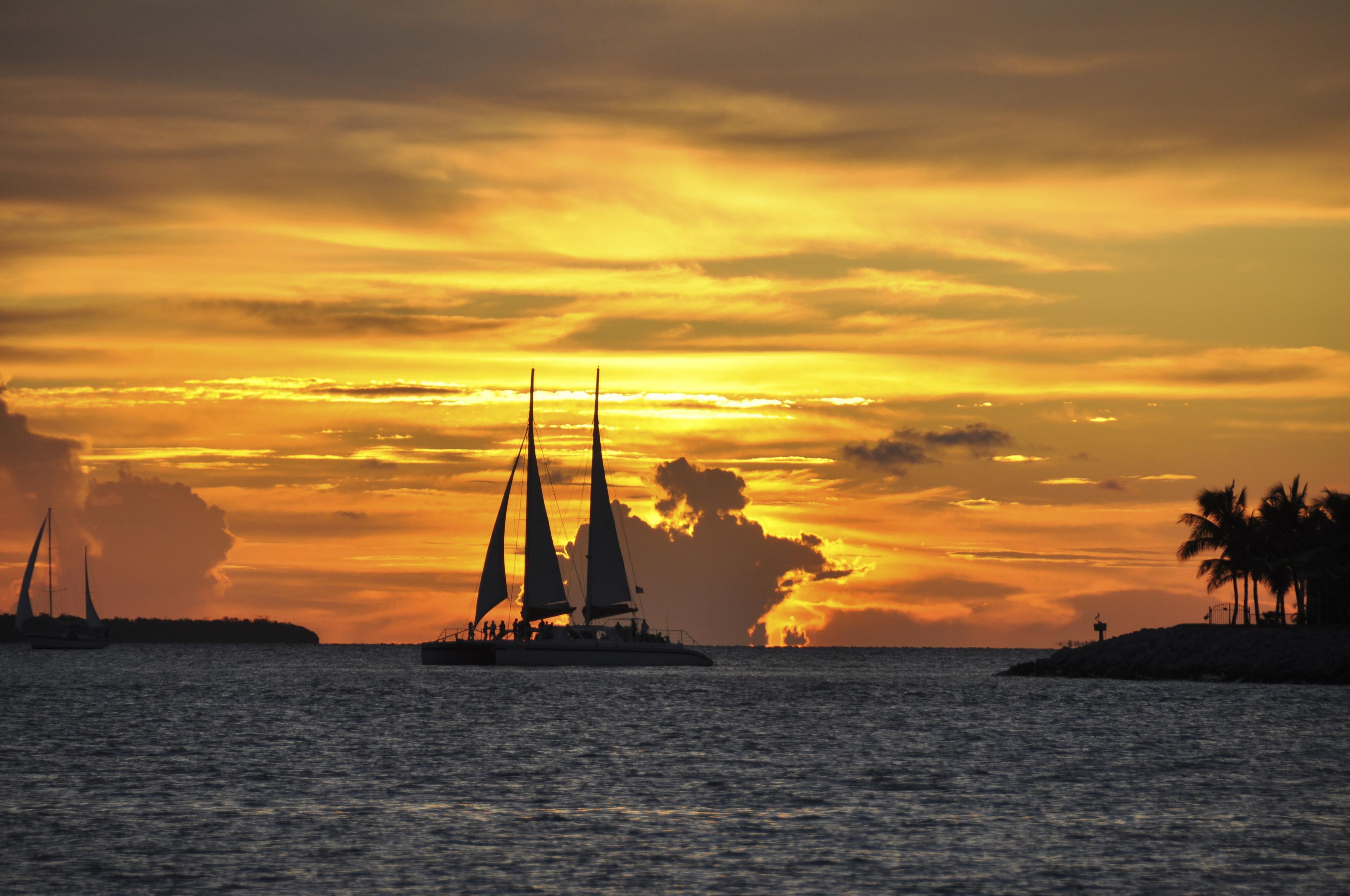 Pôr do sol sobre a água, com veleiros e uma ilha ao longe.