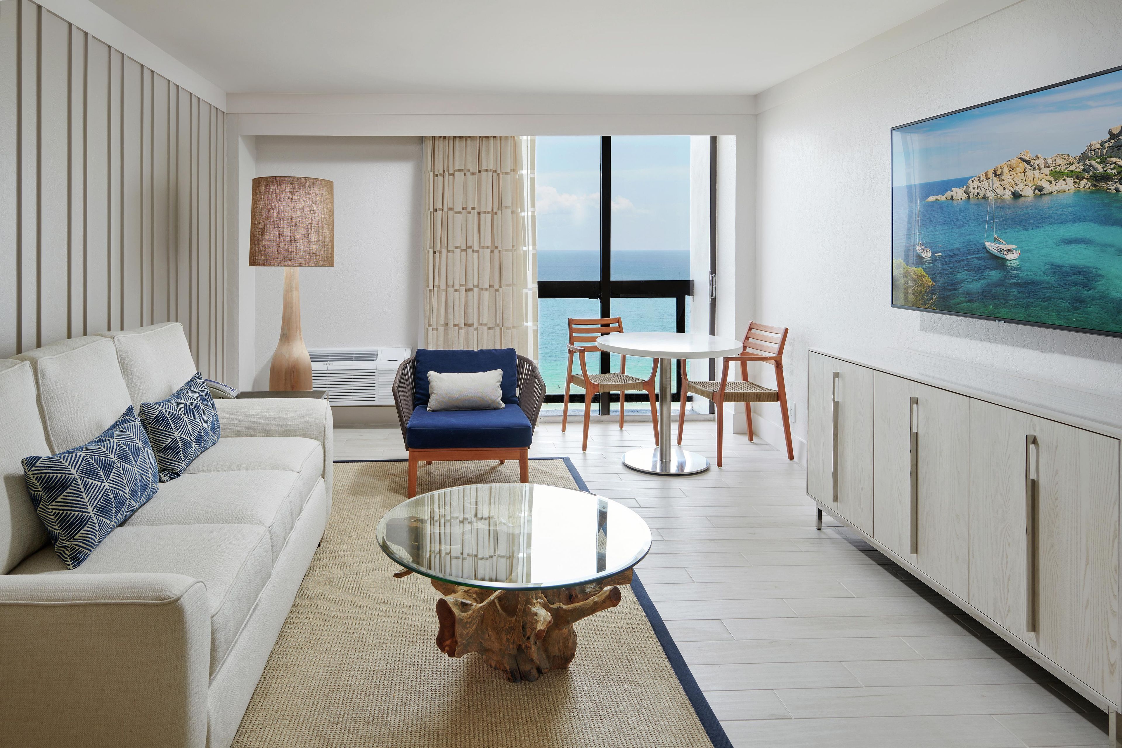 Un salon moderne avec un canapé, une table basse en verre, un coin repas avec des chaises et une grande fenêtre donnant sur la mer.
