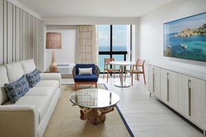 Ein modernes Wohnzimmer mit einer Couch, einem Glastisch, einem Essbereich mit Stühlen und einem großen Fenster mit Meerblick.