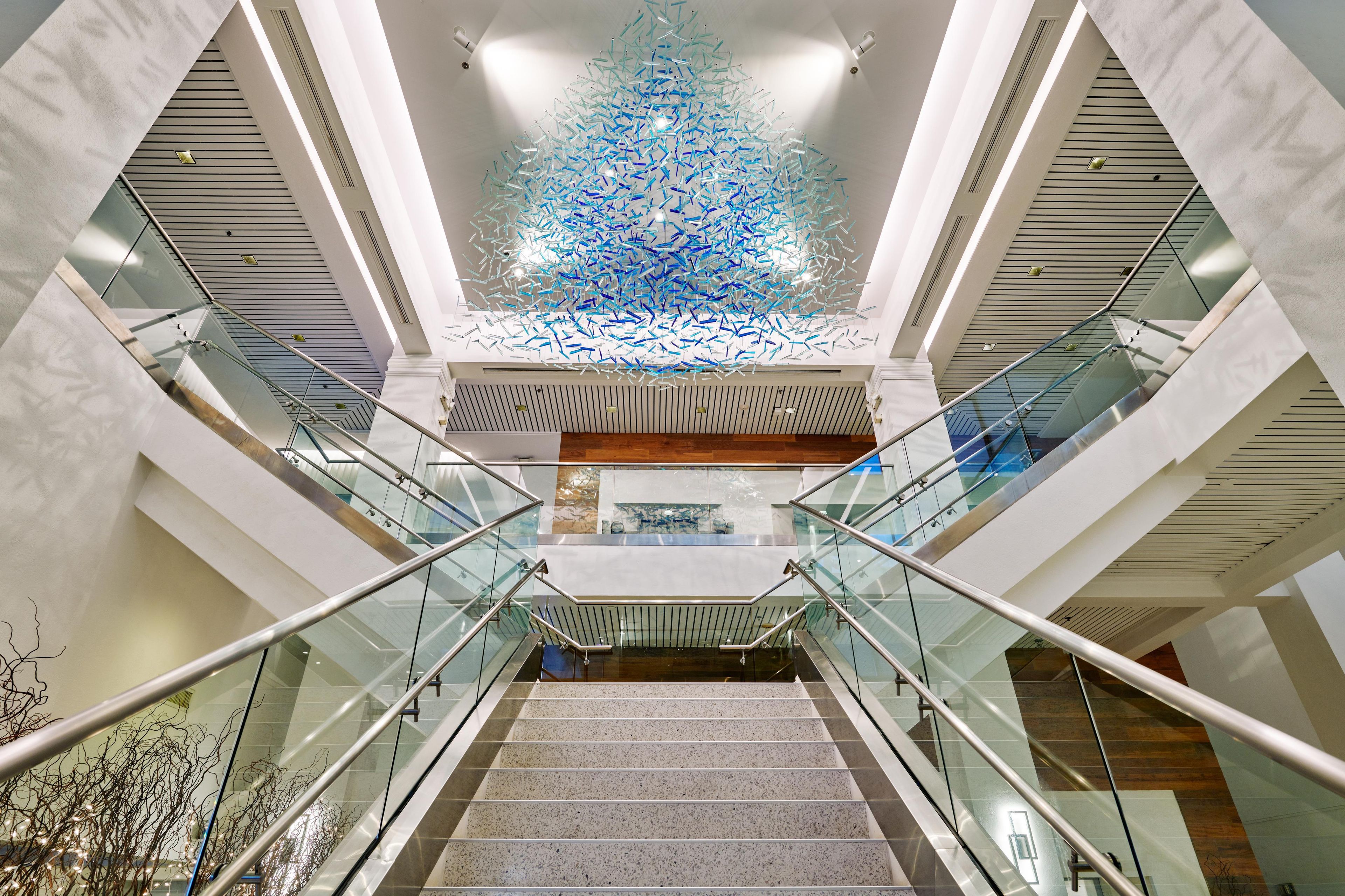 Un interior moderno con una escalera central, barandillas de vidrio e instalación decorativa en el techo.