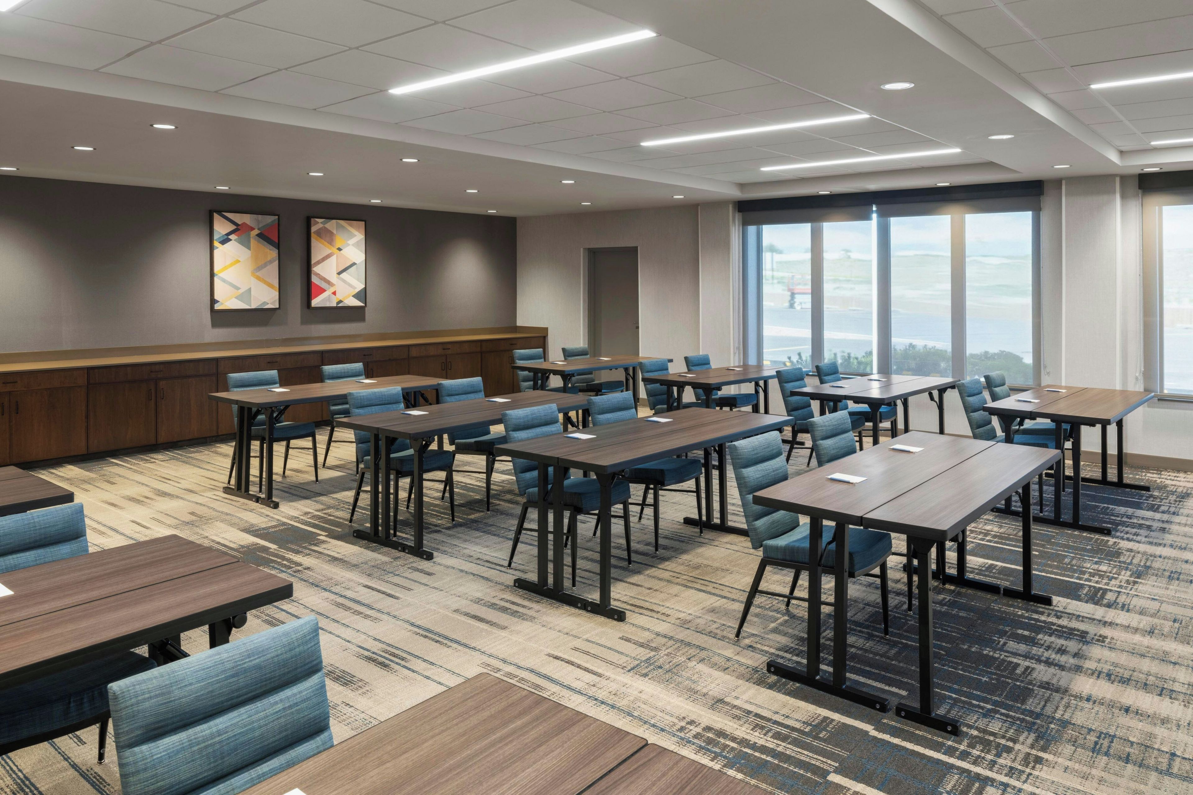 A conference room with multiple tables and chairs, a carpeted floor, and large windows offering a view.