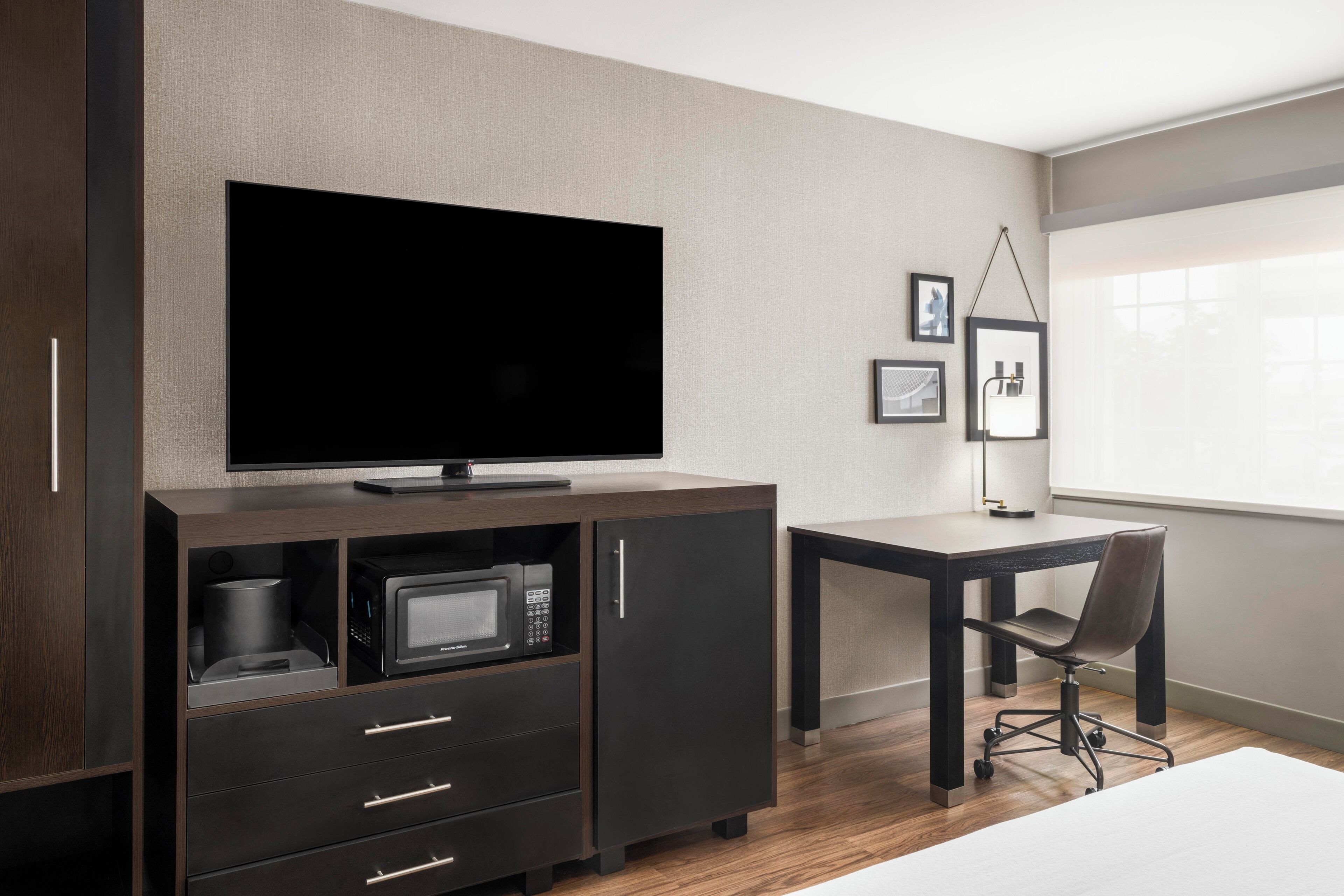 A hotel room with a flat-screen TV, a microwave, a small cabinet with drawers, a desk, a chair, and a window with blinds.