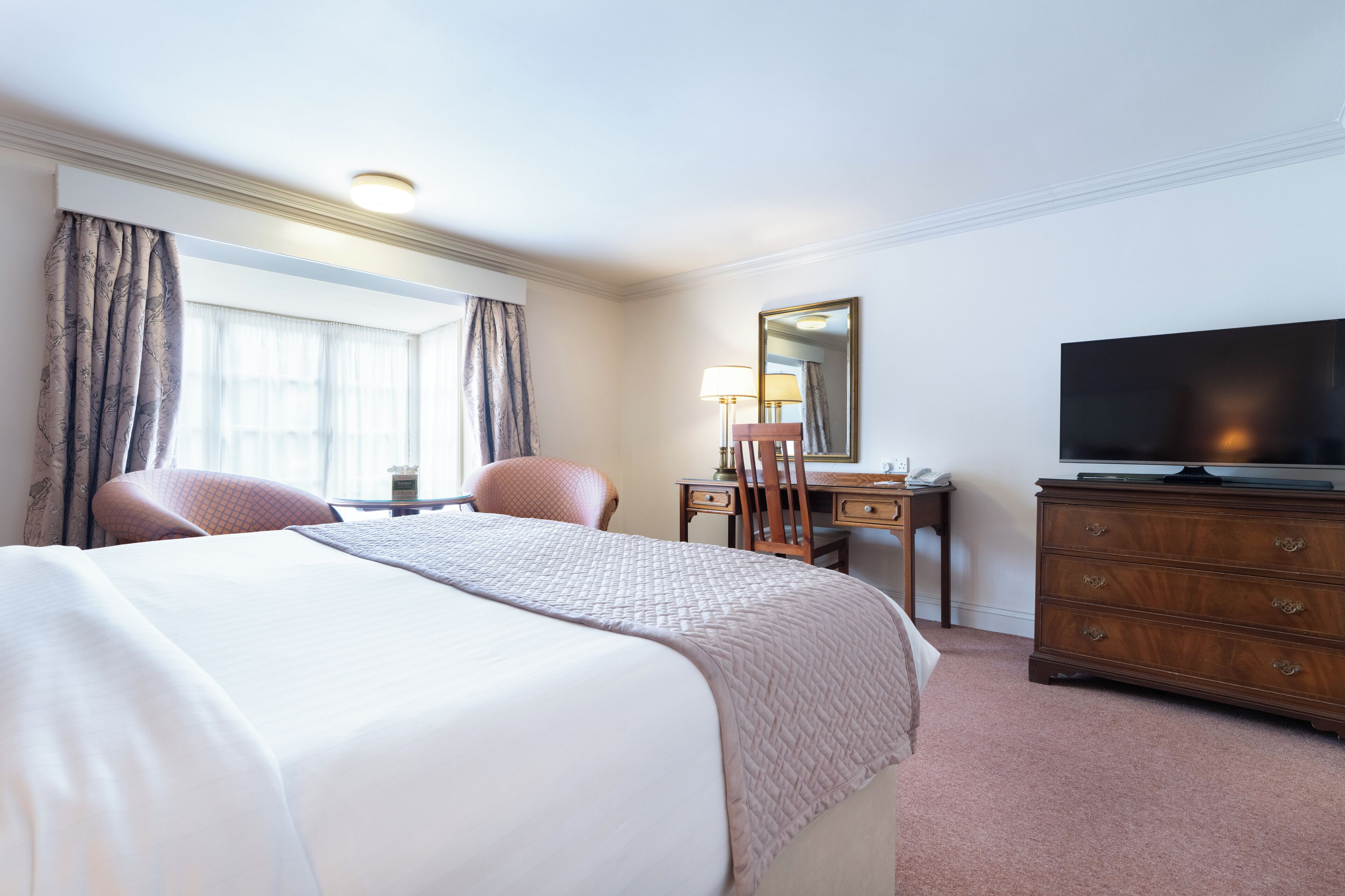 A hotel room with a large bed, a desk, a chair, a dresser, and a television.