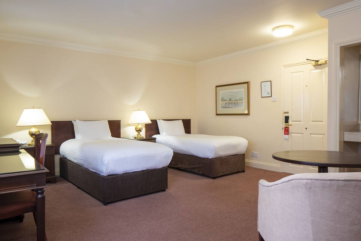 A hotel room with two beds, a desk, a chair, and a small table.