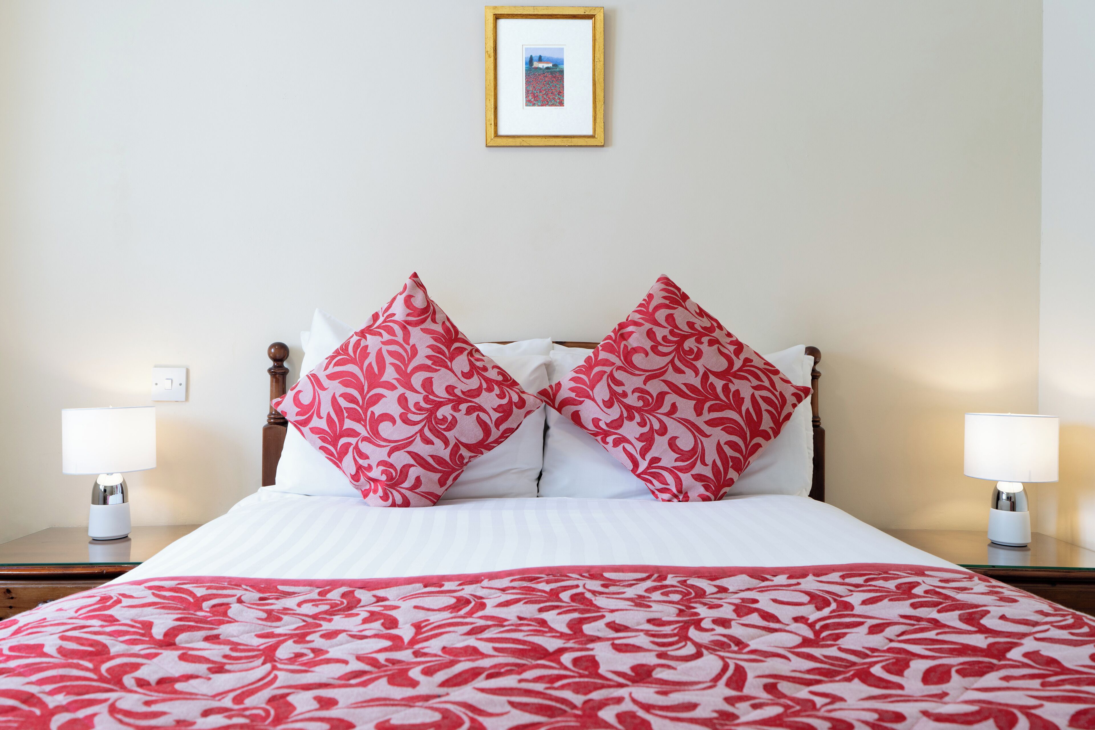 A neatly made bed with a red and white patterned bedspread and pillows, two bedside lamps, and a framed picture on the wall.