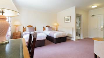 A hotel room with two beds, a desk, a chair, a lamp, and a door leading to a bathroom.
