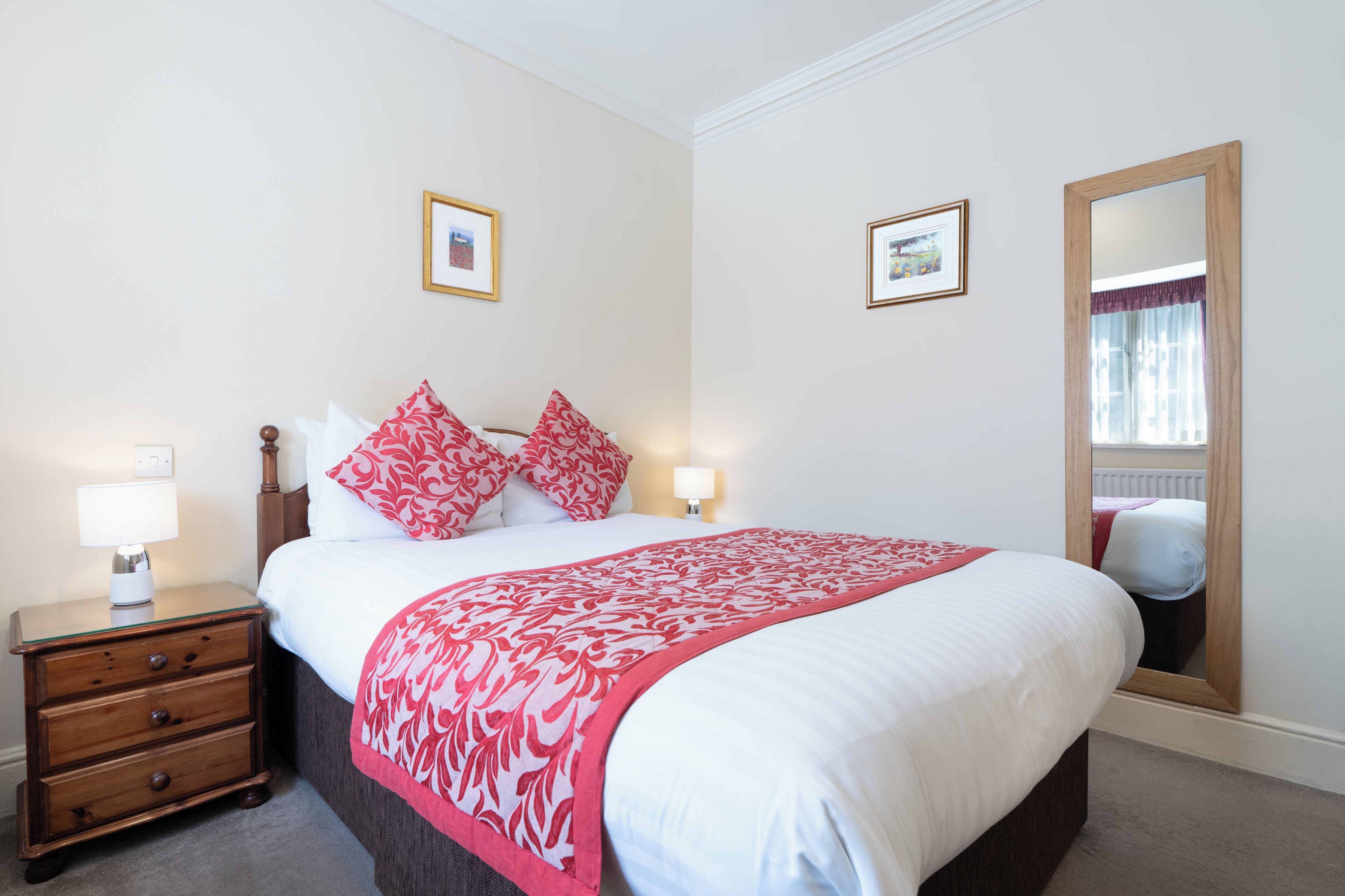 A neatly arranged bedroom with a large bed, two bedside lamps, a nightstand with drawers, and two framed pictures on the wall.