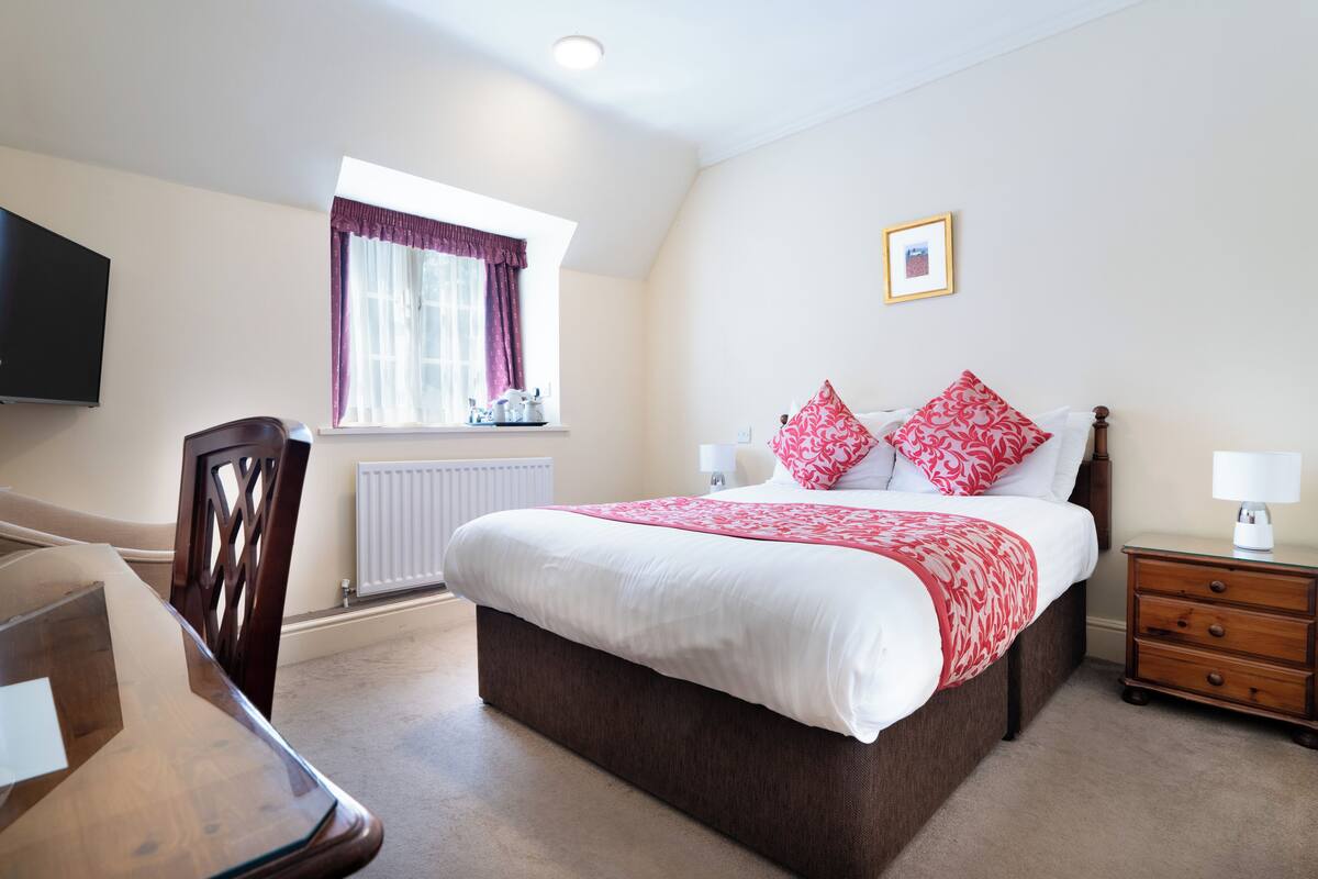 A bedroom with a large bed, a window with curtains, a bedside table with a lamp, and a TV mounted on the wall.