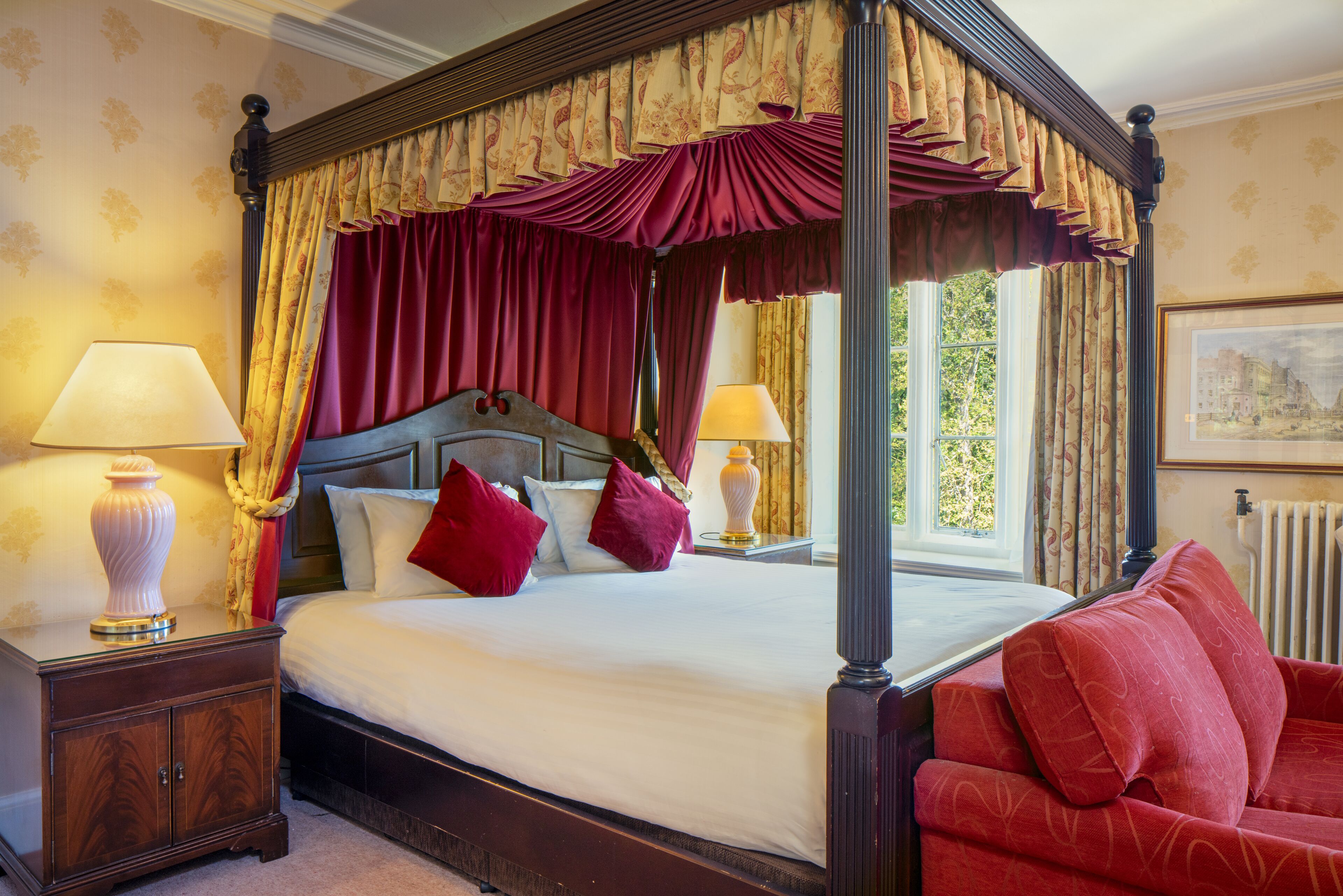 A four-poster bed with red and gold drapery, two bedside lamps, and a red sofa in a room with patterned wallpaper and a framed picture on the wall.
